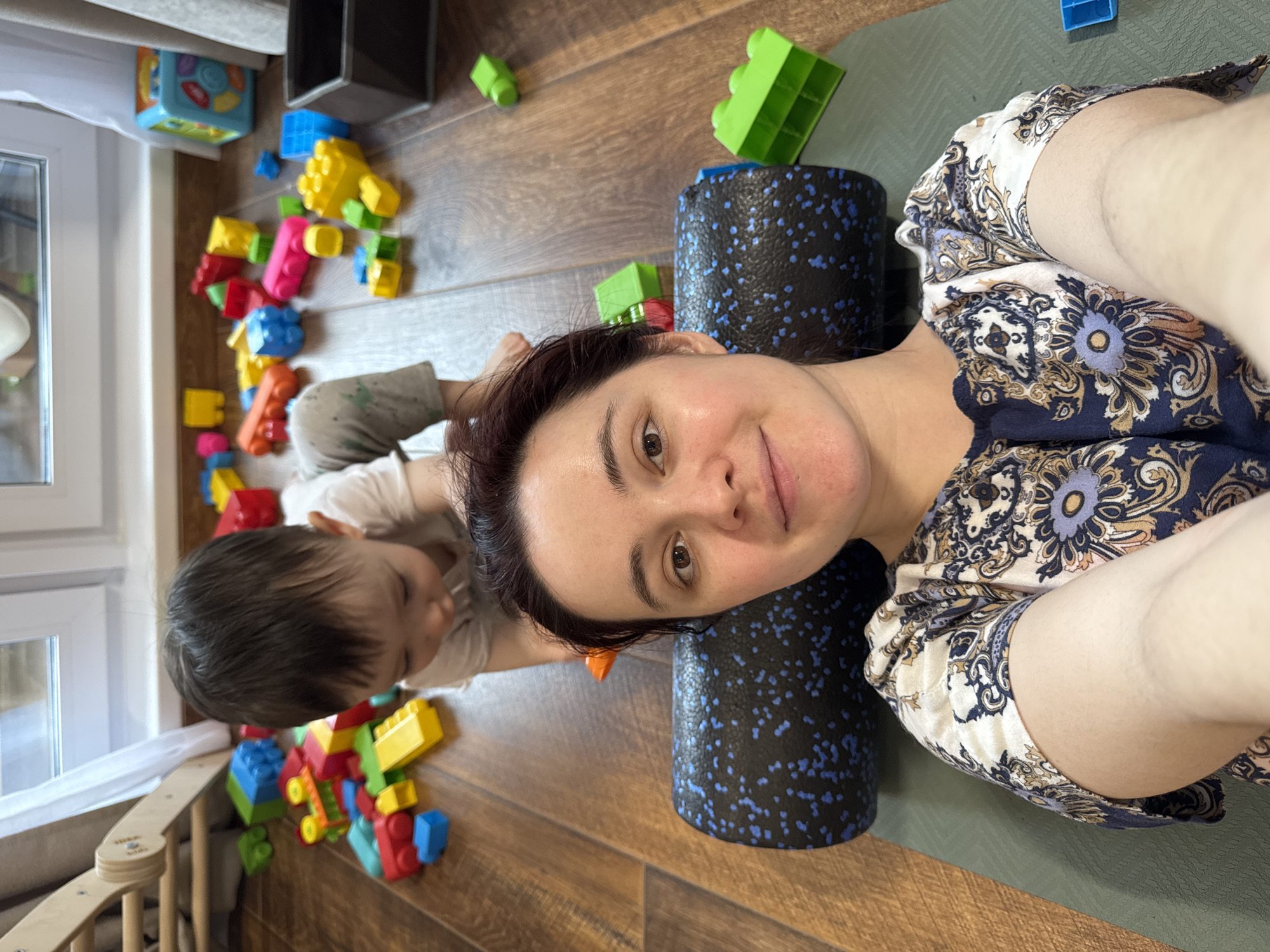 A woman is lying on a green yoga mat, using a black foam roller with blue speckles under her head. To her left is a toddler lying on the floor amidst scattered plastic building blocks and toys. The floor is wood-grain. A window is visible in the background, along with a shelf and other toy containers. The woman is wearing a patterned top. The scene appears to be indoors. There are no elements clearly indicating that the location is in Izhevsk, Russia.