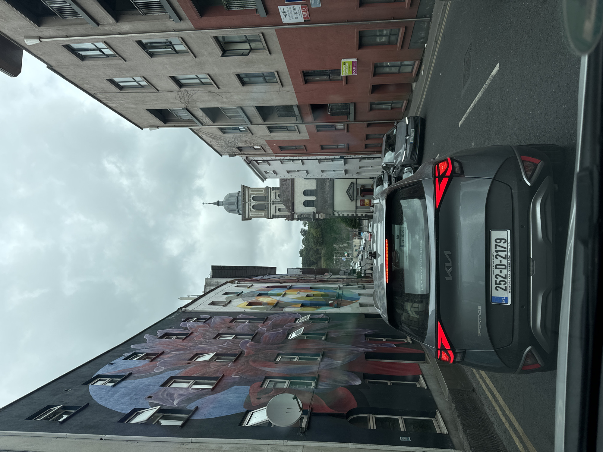The image shows a street scene in Waterford, Ireland, on a cloudy day. A gray Kia Sportage is in the foreground, facing away from the camera, with its brake lights illuminated. The car's license plate reads "252-D-2179". To the left of the Kia is a building adorned with a large, colorful mural of abstract floral shapes. Several windows on this building are open. To the right of the Kia, the street continues, lined with apartment buildings. The building directly to the right of the Kia is painted in shades of beige and reddish-brown, with several balconies and windows. Further down the street, on the left side, a building with a dome and tower, resembling a church or public building, is visible. Traffic is present on the street, with other cars and a traffic light visible in the distance. The street is paved with asphalt, and there are yellow lines marking the lanes. The environment appears to be a mix of residential and commercial buildings.