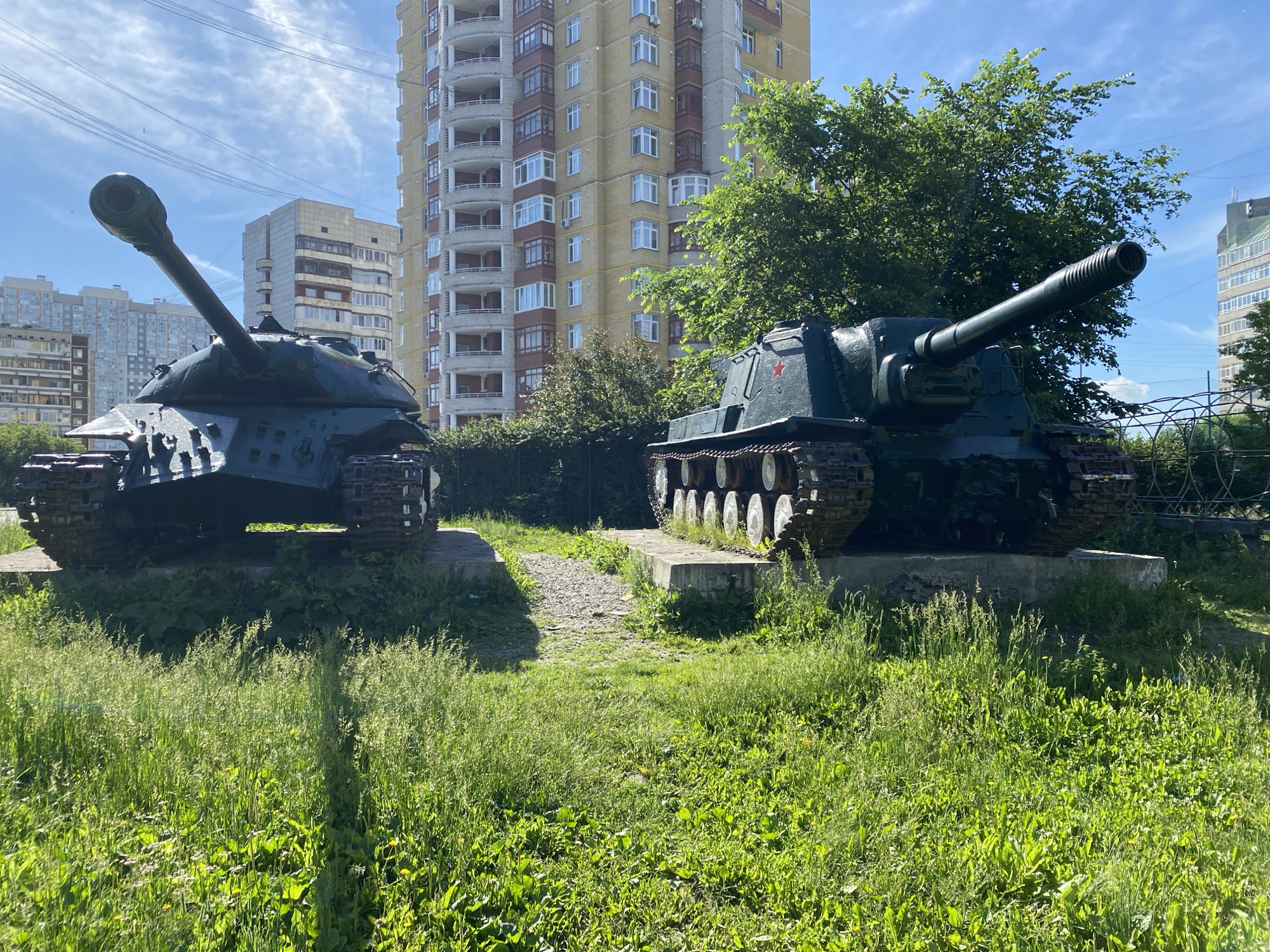 The image depicts two decommissioned Soviet tanks, likely IS-3s, displayed on concrete pedestals amidst lush green grass and trees. In the background, modern high-rise apartment buildings with balconies are visible against a bright blue sky with scattered clouds. The tanks are positioned in an outdoor urban setting, possibly a memorial or a park, within the city of Yekaterinburg, Russia. The scene is characterized by daylight and appears peaceful, with no people present. The tanks are dark green and show signs of age, indicating they are historical exhibits.