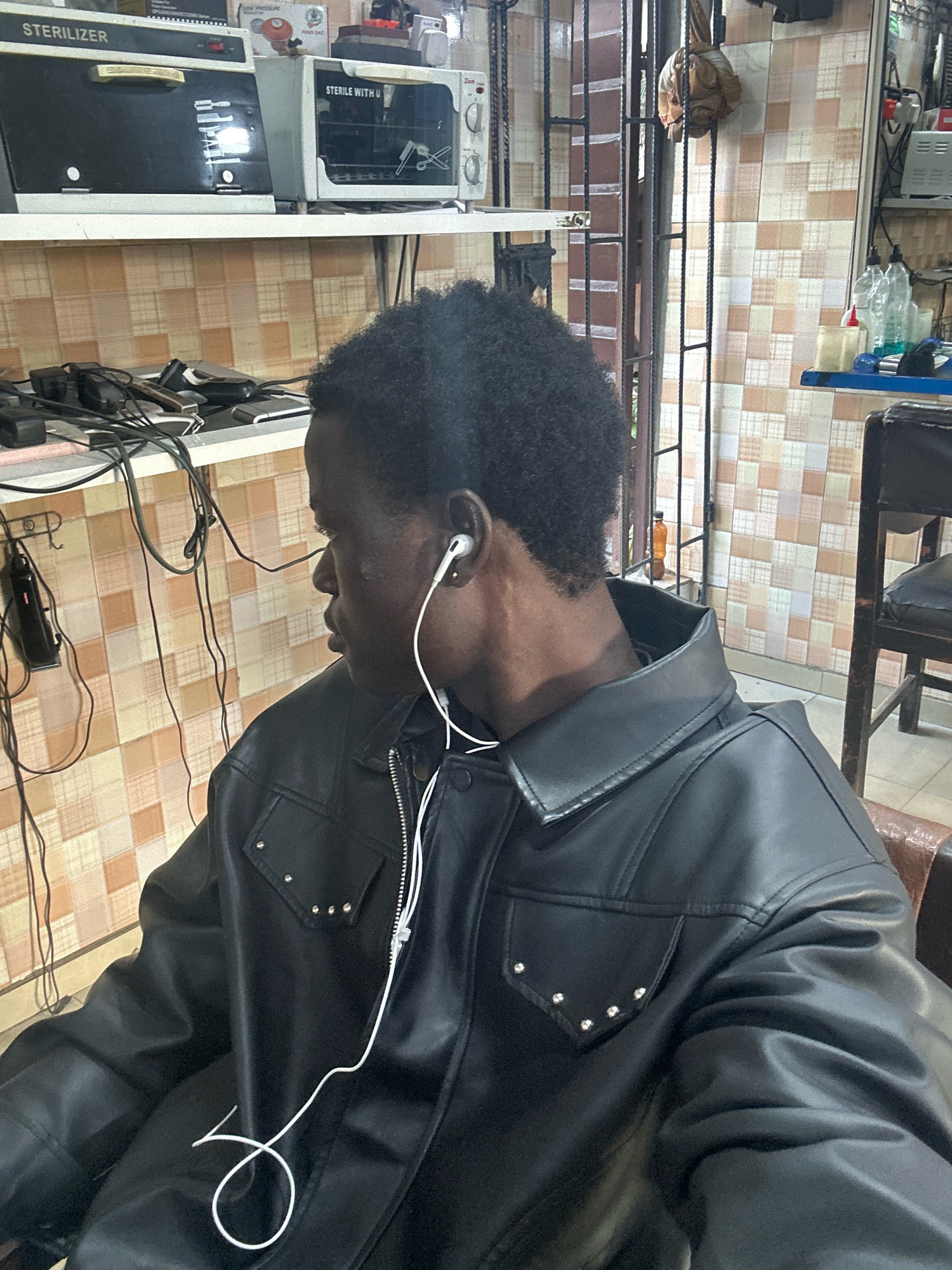 The image shows a young Black male wearing a black leather jacket and white earbuds, seated in what appears to be a barber shop in Badagry, Nigeria. He is looking to his left, away from the camera. The background shows shelves with various items, including sterilizers, electrical equipment, and bottles. The walls are tiled in a checkered pattern of cream and light brown. A metal grate structure is visible on the right side of the image, and a chair with a leather seat is partially visible. The lighting suggests an indoor environment, possibly with artificial light sources.
