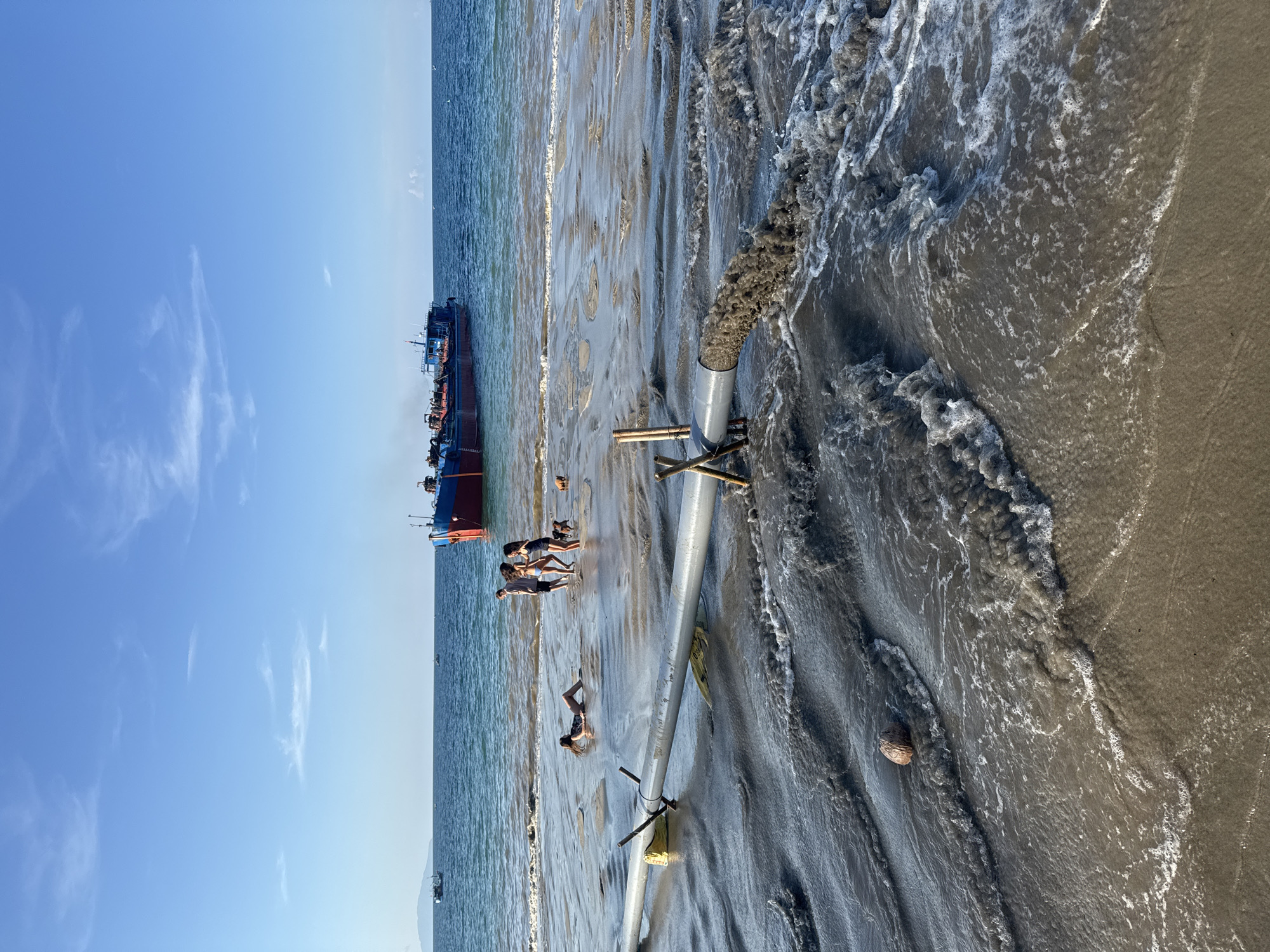 A horizontal shot depicts a beach scene with a large, red cargo ship in the distance on the left, and a wide, silver pipe running along the beach, its open end spewing dark sediment into the ocean on the right. Several individuals are wading in the water near the ship and pipe, with one lying in the shallow water. The sky is blue with scattered clouds, and the beach is wet with visible wave patterns. A coconut is seen floating near the waves. The location is the beach in Đà Nẵng, Vietnam.
