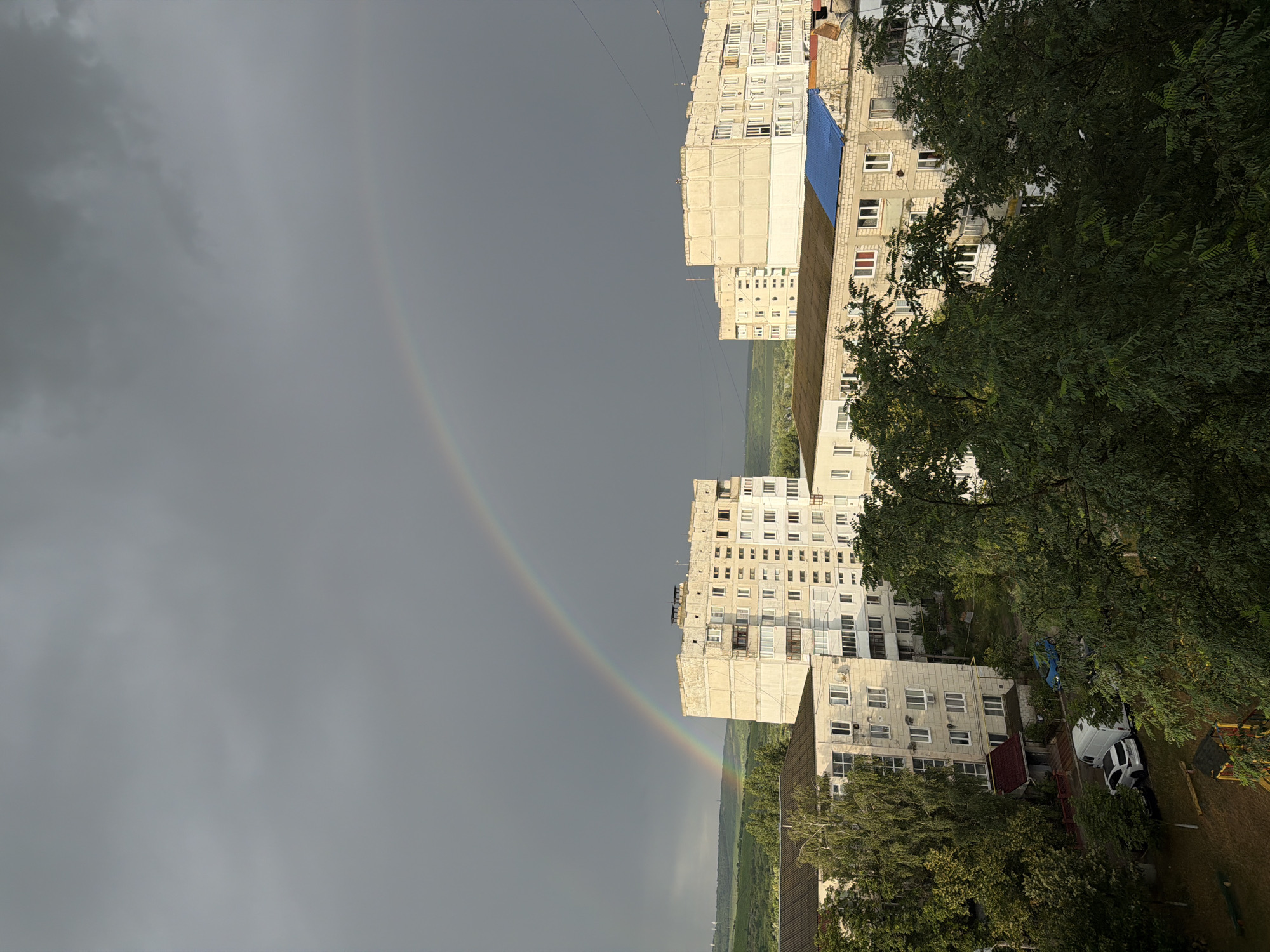 The image depicts an urban landscape in Strășeni, Moldova, under a dramatic sky. A prominent rainbow arcs across a heavily overcast, grey sky, hinting at recent or ongoing precipitation. In the foreground, several apartment buildings dominate the scene, characterized by their multi-story, block-like architecture typical of post-Soviet urban planning. One building in the middle ground is a light beige color with numerous windows and balconies, showing signs of wear. To its right, another similar building stands, also exhibiting utilitarian design. In the lower part of the image, lush green trees partially obscure the view of the buildings and a residential area, where a white van and a dark car are parked on a paved area. A small playground structure is visible in the bottom left corner. The background reveals rolling green hills, suggesting the proximity of rural or natural landscapes to the city. The overall impression is one of a somewhat weathered urban environment set against a striking natural atmospheric phenomenon.