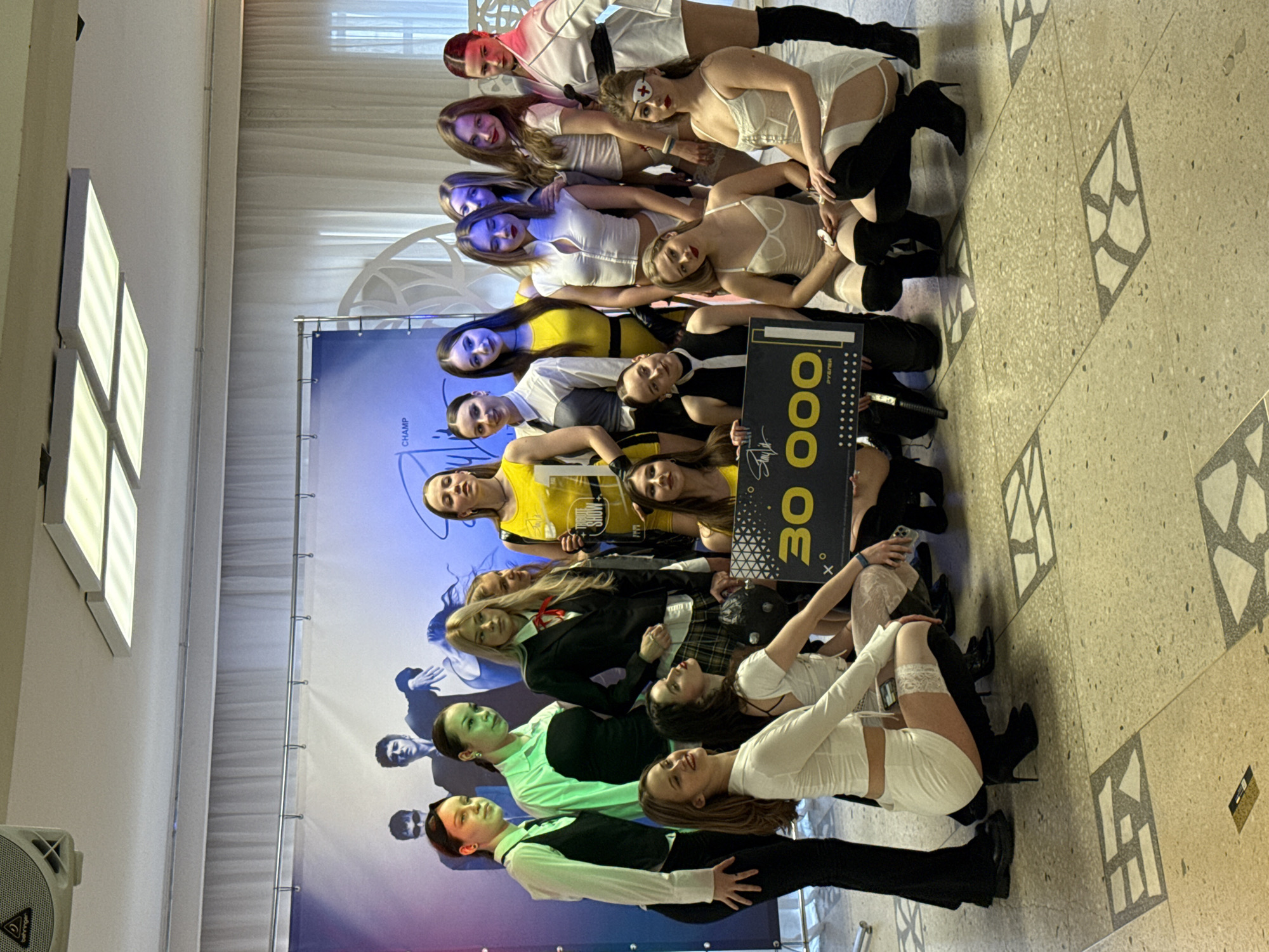 A group of approximately 15 young women are posing for a photo in what appears to be an indoor event hall. The women are dressed in varied, somewhat provocative outfits, with a mix of formal and playful attire. They are arranged in a stacked formation. The group is holding a large sign with the number "30 000" on it, likely representing a prize or reward. Behind them is a backdrop banner with an image of people and a logo. The location is suggested to be in Perm, Russia, but there is no immediate visual confirmation in the image. The environment is a polished interior, with patterned flooring and artificial lighting visible. The interaction is a celebratory group pose, suggesting a successful event.