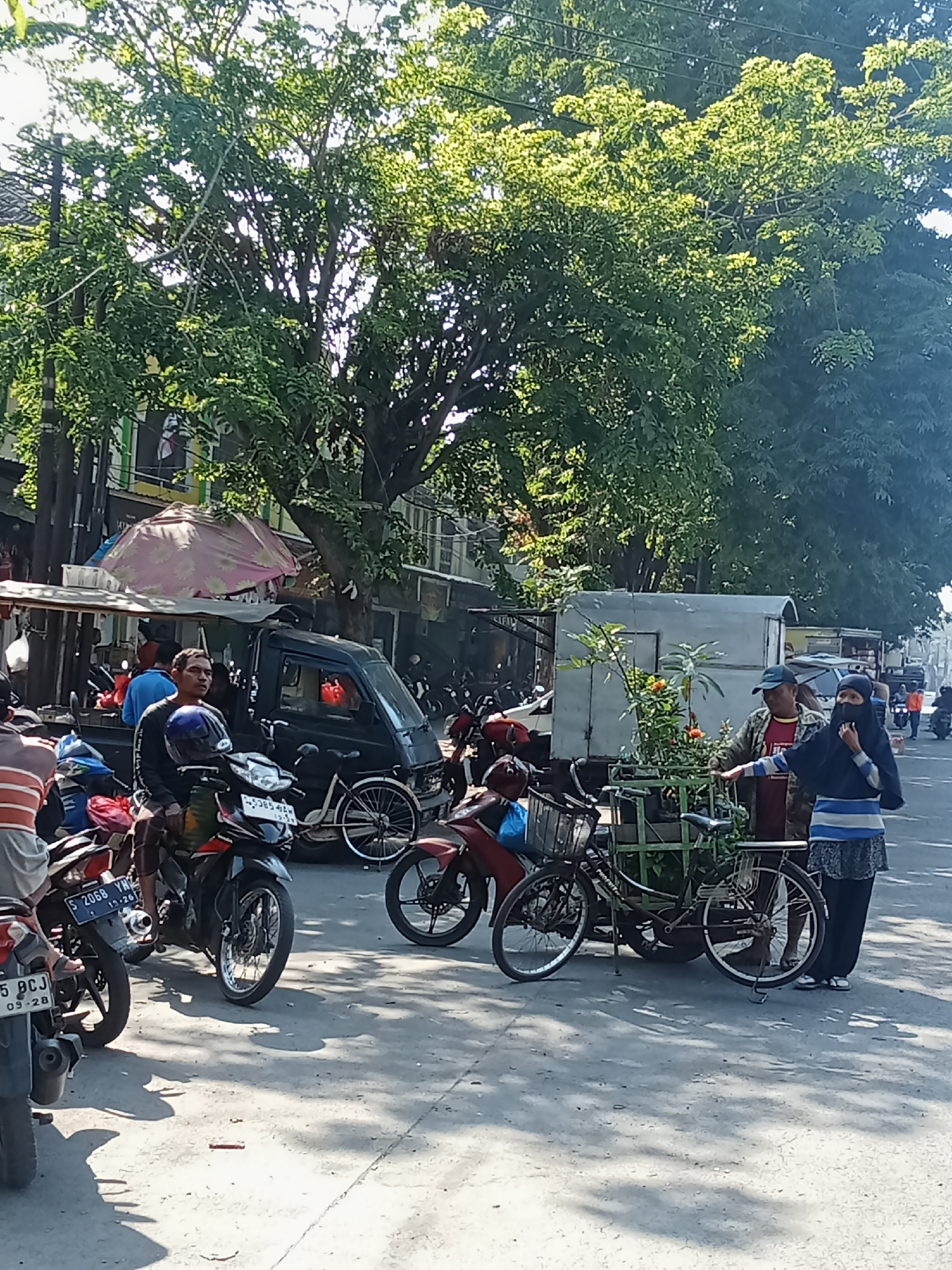 The image depicts a street scene in Peterongan, Indonesia. In the foreground, several motorcycles are parked and in motion. A man wearing a blue shirt is seated on a black motorcycle. To his right, a black pickup truck is parked. Further back, a woman in a dark blue hijab and a striped shirt is standing next to a man wearing a checkered jacket and a red shirt. They are near a bicycle with plants in a green holder. The background shows buildings, trees, and a bright sky, indicating it is daytime. The street appears to be paved and is filled with sunlight and shadows.