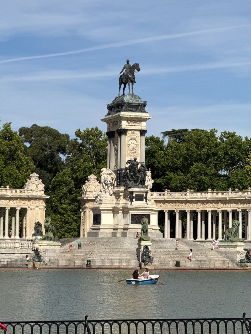 The image captures a vibrant daytime scene at the Monument to Alfonso XII in El Retiro Park, Madrid, Spain. The central focus is a grand, elaborate monument made of light-colored stone. At its pinnacle stands a large equestrian bronze statue of a man, presumably King Alfonso XII, mounted on a horse. Below him, the monument's pedestal is adorned with classical columns, intricate carvings, and additional smaller sculptures. Behind this central structure, a sweeping semi-circular colonnade with numerous white columns and more sculptures completes the grand architectural display.
In front of the monument stretches a calm body of water, a lake or pond, where several small rowboats are visible. In the foreground, a light blue rowboat, clearly marked with the number "18" on its side, is occupied by two individuals who appear to be rowing. Numerous people are scattered across the scene; some are seated on the broad stone steps leading up to the monument, while others walk along the monument's base or near the colonnade.
Lush green trees frame the monument on both sides and in the background, providing a natural, verdant setting. The sky above is a clear, bright blue with a few faint wispy clouds and a single contrail. The bright sunlight indicates it is likely midday or early afternoon. A black metal fence runs across the bottom of the frame, separating the viewer from the water. The overall atmosphere suggests a pleasant day with people enjoying leisurely activities in a historical park setting.