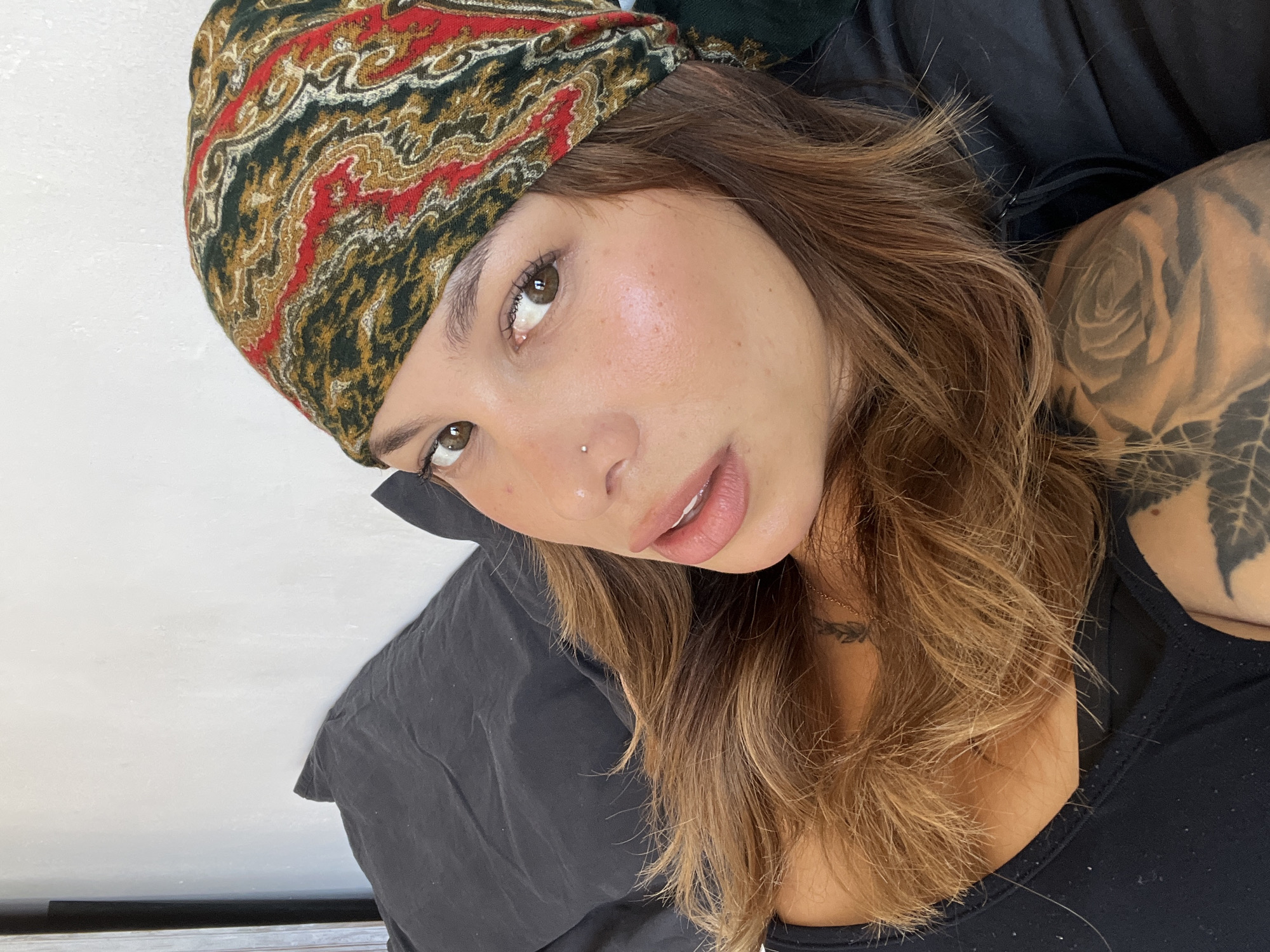 The image shows a medium shot of a woman lying down, resting her head on a black pillow. She is wearing a colorful bandana on her head and a black top. Her hair is brown and falls over her shoulders. She has a nose piercing and a tattoo on her neck. Part of another tattoo is visible on her arm. The background consists of a white wall and a black pillow. The scene appears to be indoors, possibly in a bedroom. There is no indication of the specific location within Barcelona, Spain.