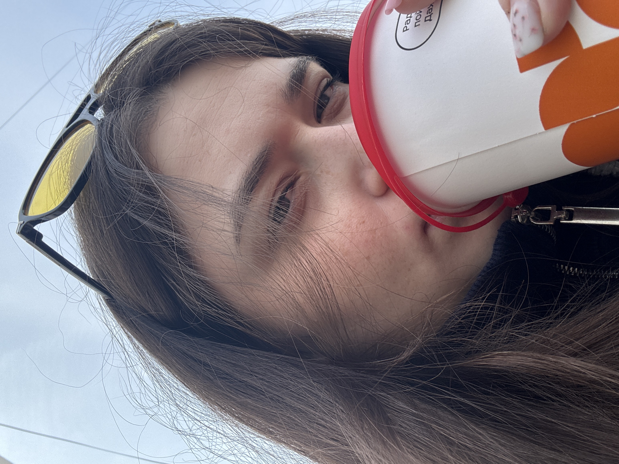 A woman with brown hair is drinking from a white and orange cup with a red lid. She is wearing sunglasses. The location is Chaikovsky, Russia, as indicated by the Russian text on the cup. The background is a light sky.