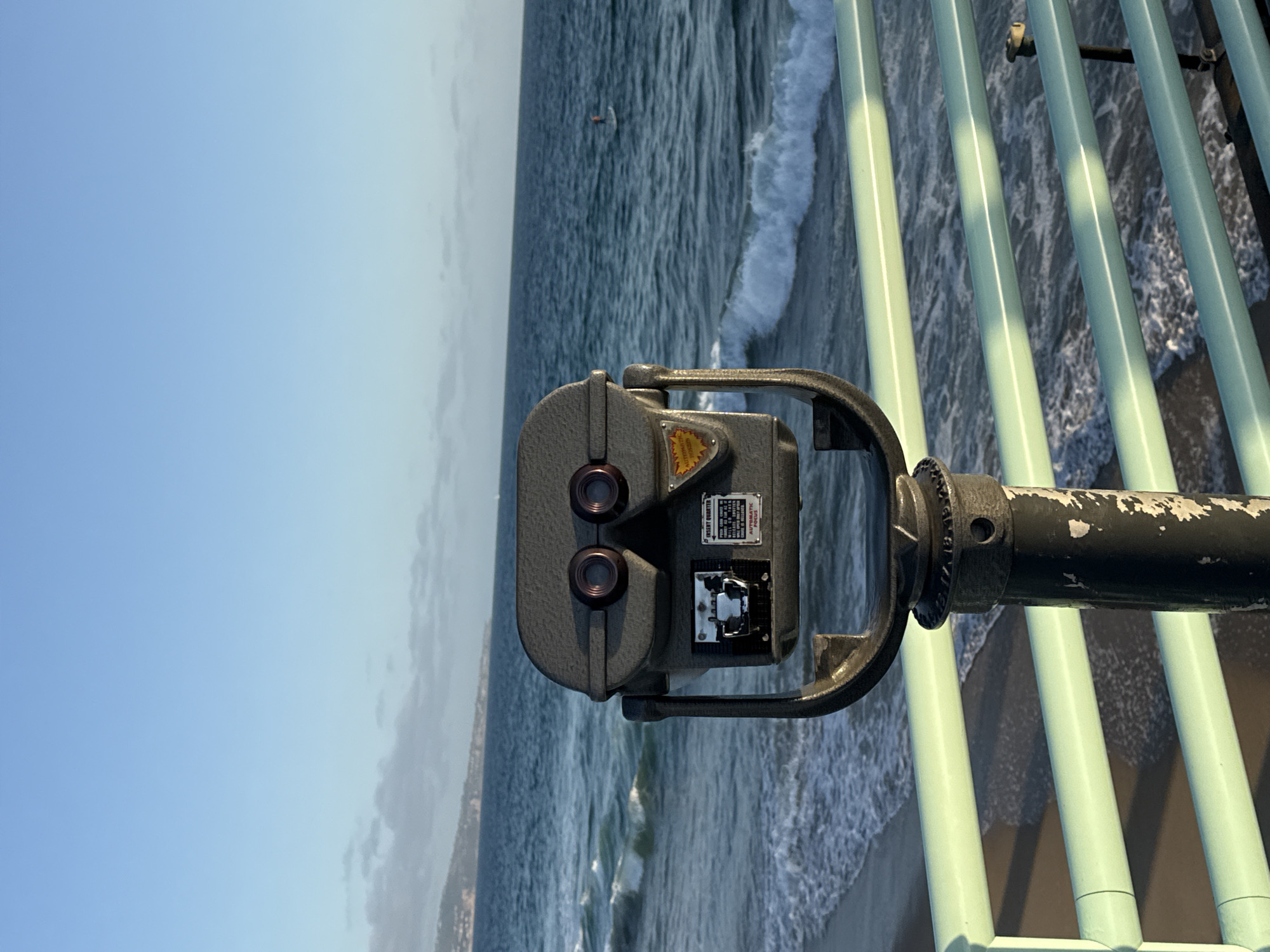 This is a medium shot, vertical photograph taken from a pier in Manhattan Beach, California, USA. In the foreground, a weathered, dark grey, coin-operated binocular viewer stands on a black pole. The viewer has two eyepieces and a slot for inserting coins. A sign on the viewer reads "INSERT QUARTER" and "AUTOMATIC FOCUS." The viewer is positioned in front of a pale green metal railing, with three horizontal bars visible.
Beyond the railing, the ocean stretches to the horizon. Gentle waves with white foam are breaking on a sandy beach, which is visible in the lower left portion of the image. The water is a deep blue, with lighter blue and white accents from the waves. In the distance, a silhouette of a person on a paddleboard is visible in the ocean. Further in the background, a hazy coastline with hills can be seen under a clear, pale blue sky. The lighting suggests it is late afternoon or early evening, with the sun casting shadows from the railing onto the beach.