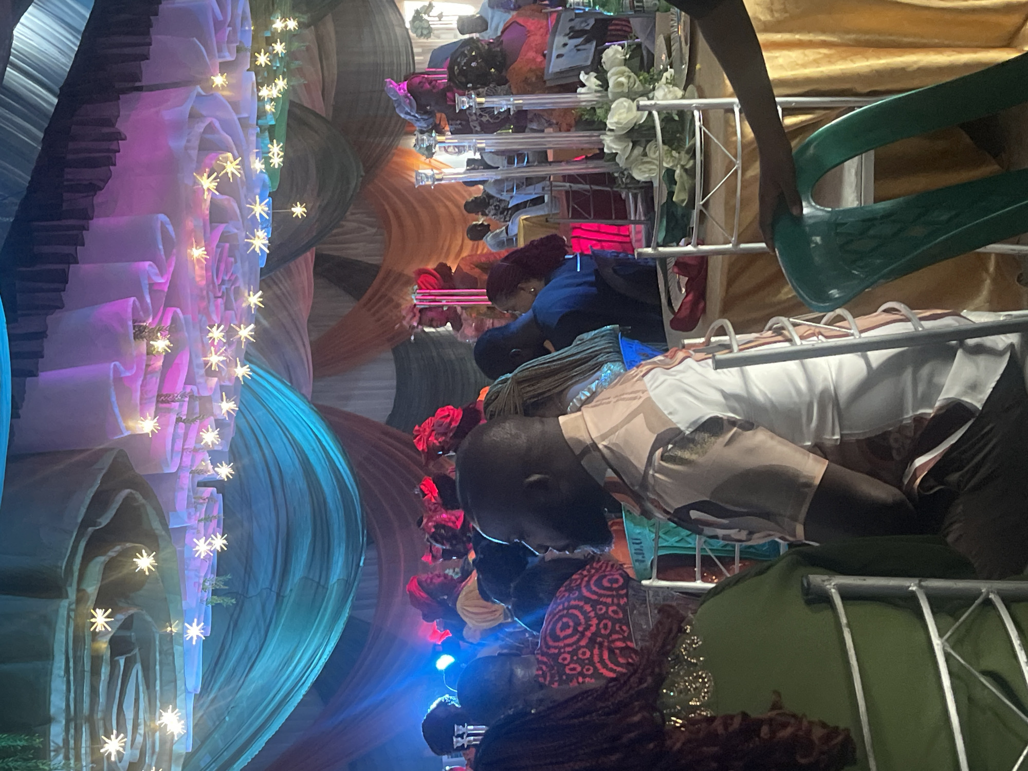 The image captures an indoor celebratory event, likely taking place in the evening or night, given the reliance on artificial lighting. The scene is festive and vibrant, with numerous decorative elements.
The setting appears to be a large hall or tent, intricately adorned with draped fabrics in various colors, including shades of pink, blue, green, brown, and orange. These fabrics create a wavy, textured ceiling or wall effect. Numerous star-shaped string lights are strung across the draped materials, casting a warm, sparkling glow and enhancing the festive atmosphere.
A group of people are visible, mostly from behind or in profile, seated at tables. They appear to be engaged in a social gathering or meal. In the foreground, a person wearing a white t-shirt with a brown and tan camouflage pattern is seated. Further back, another individual wears a dark blue top. Many of the attendees have dark hair, and some wear head coverings or intricate hairstyles. The tables are set up with white metal frames, some covered with golden fabric, and feature white floral arrangements. A person's hand is visible in the upper right, holding a large, curved green plastic object, possibly a part of the serving equipment or a decorative element.
The overall impression is one of a lively and well-decorated event, consistent with a social celebration or banquet in Jos, Nigeria. No discernible text is visible in the image.