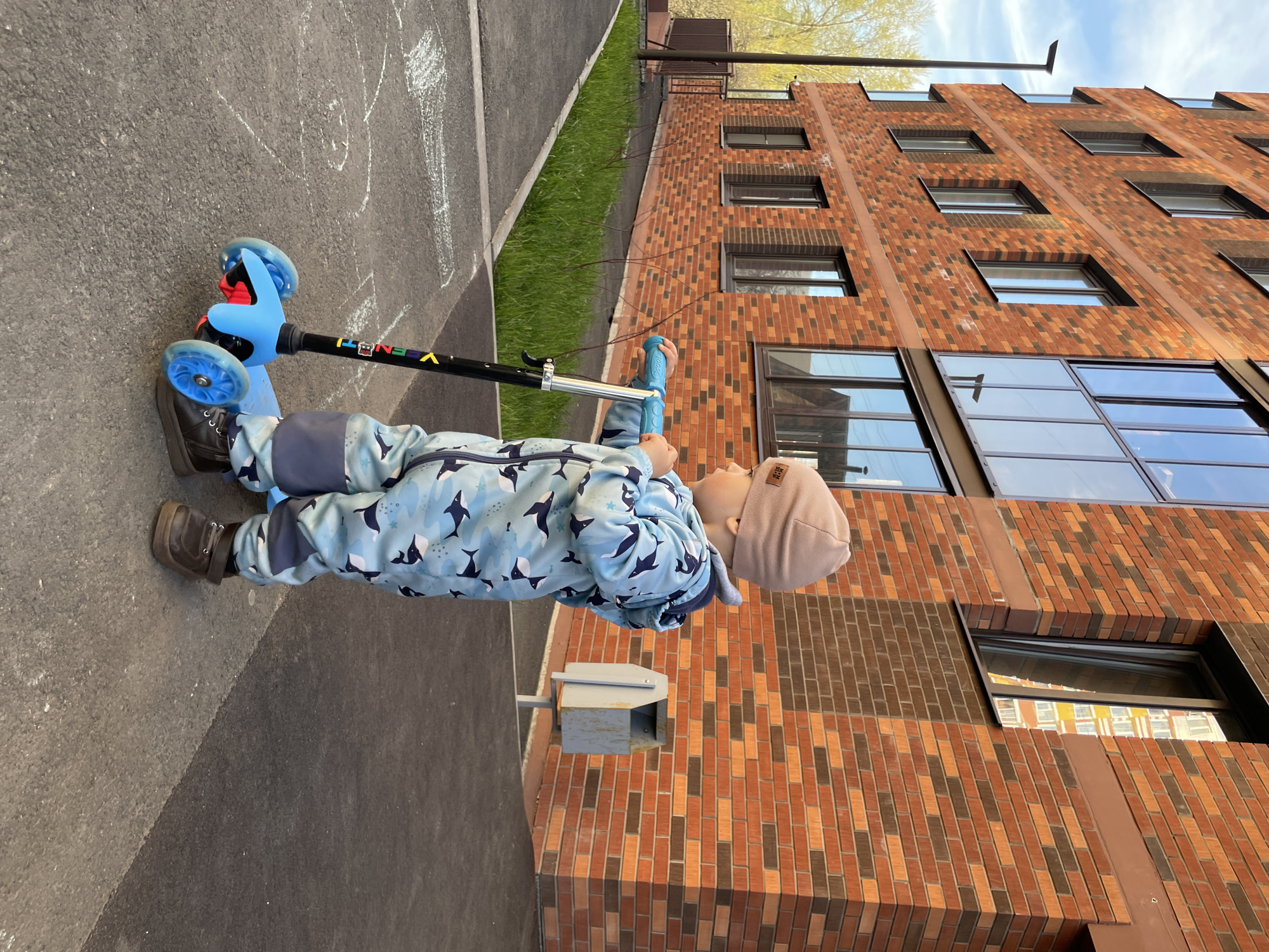 A child in a light blue patterned snowsuit and a tan beanie is riding a blue and black scooter on a grey concrete surface next to a brick building. The building has a combination of red and dark brown bricks and multiple windows. A strip of green grass is visible between the concrete and the building. A utility box is mounted on the building. The sky is blue with some clouds visible. The location is Izhevsk, Russia.