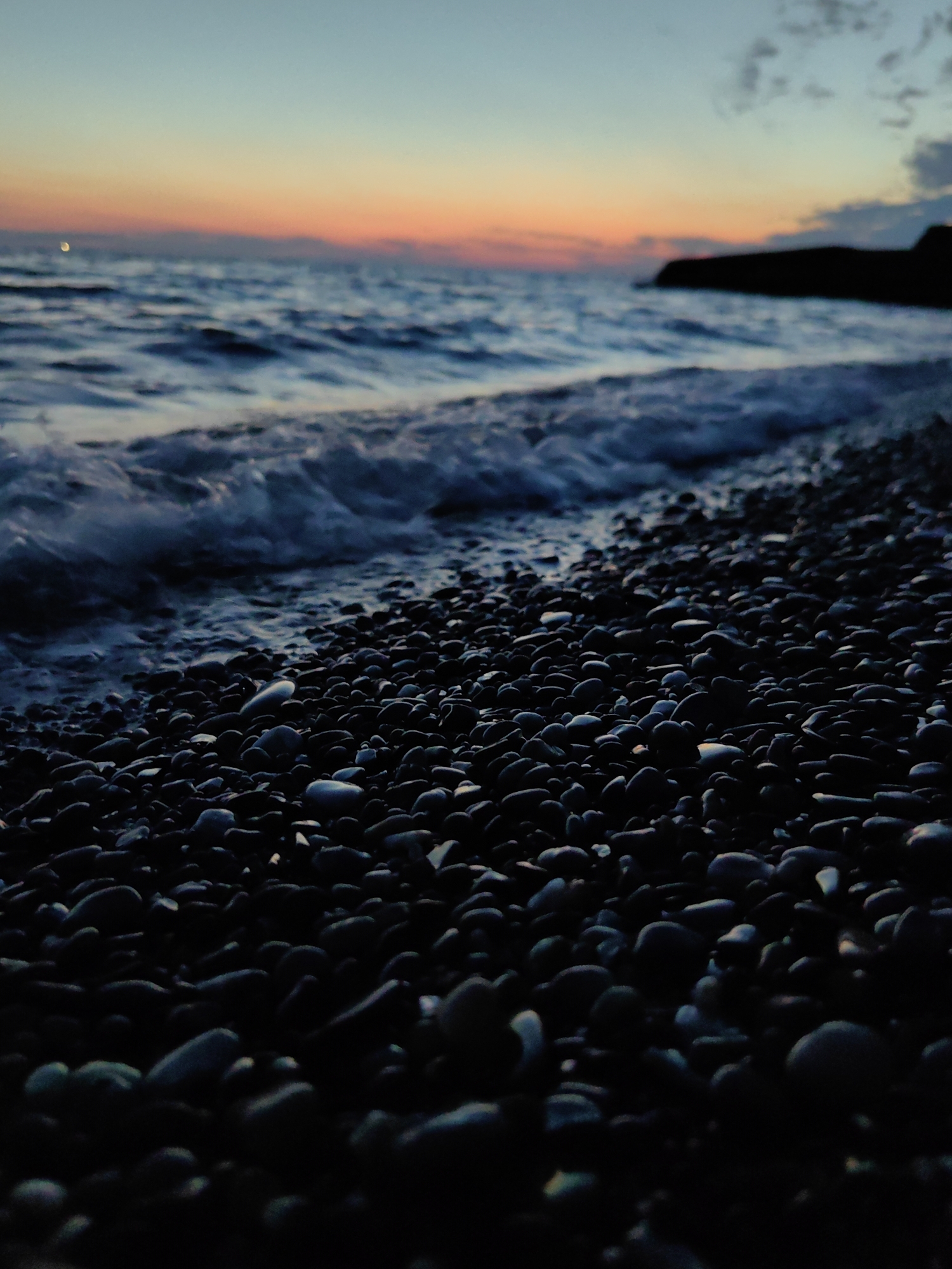 A low-angle, close-up shot captures a rocky shoreline at dusk. The foreground is dominated by dark, smooth pebbles that fill the frame, some reflecting faint light. Beyond the pebbles, the sea is a deep blue with frothy white waves lapping at the shore. The horizon shows a blend of warm orange and pink hues from the setting sun, transitioning to a pale blue sky with scattered clouds. A dark, silhouetted headland juts out into the sea on the right side of the image. A faint crescent moon is visible in the upper left portion of the sky. The scene suggests a tranquil evening by the Black Sea, likely in Sochi, Russia, given the rocky coast and twilight atmosphere. There are no people or discernible actions depicted.