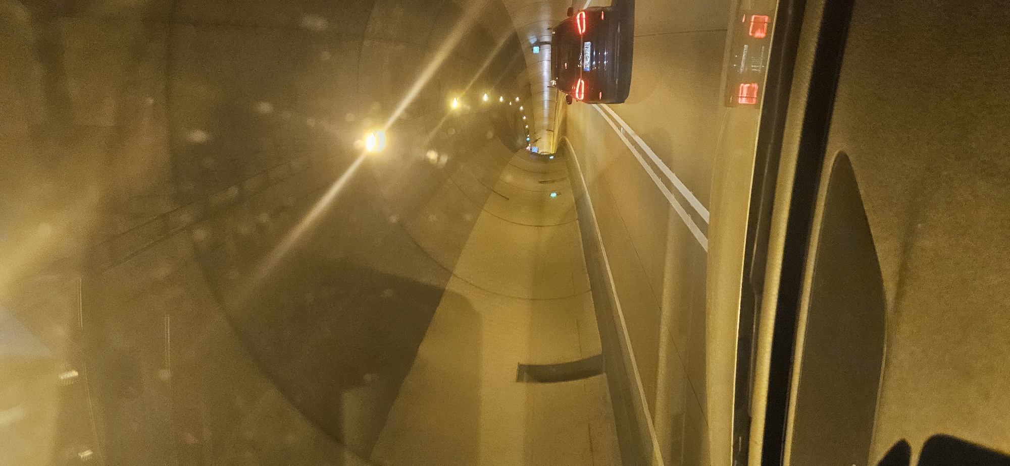 A dark-colored SUV with a Polish license plate is traveling through a tunnel in Čadca, Slovakia. The tunnel is illuminated by overhead lights, casting a warm, yellow glow on the road and walls. The road is marked with white double lines, indicating two lanes of traffic. The SUV is in the right lane, and its brake lights are illuminated. The image is taken from the perspective of another vehicle, with the dashboard and windshield visible in the foreground. The tunnel walls are curved and appear to be made of concrete. There are no other vehicles or people visible in the image.