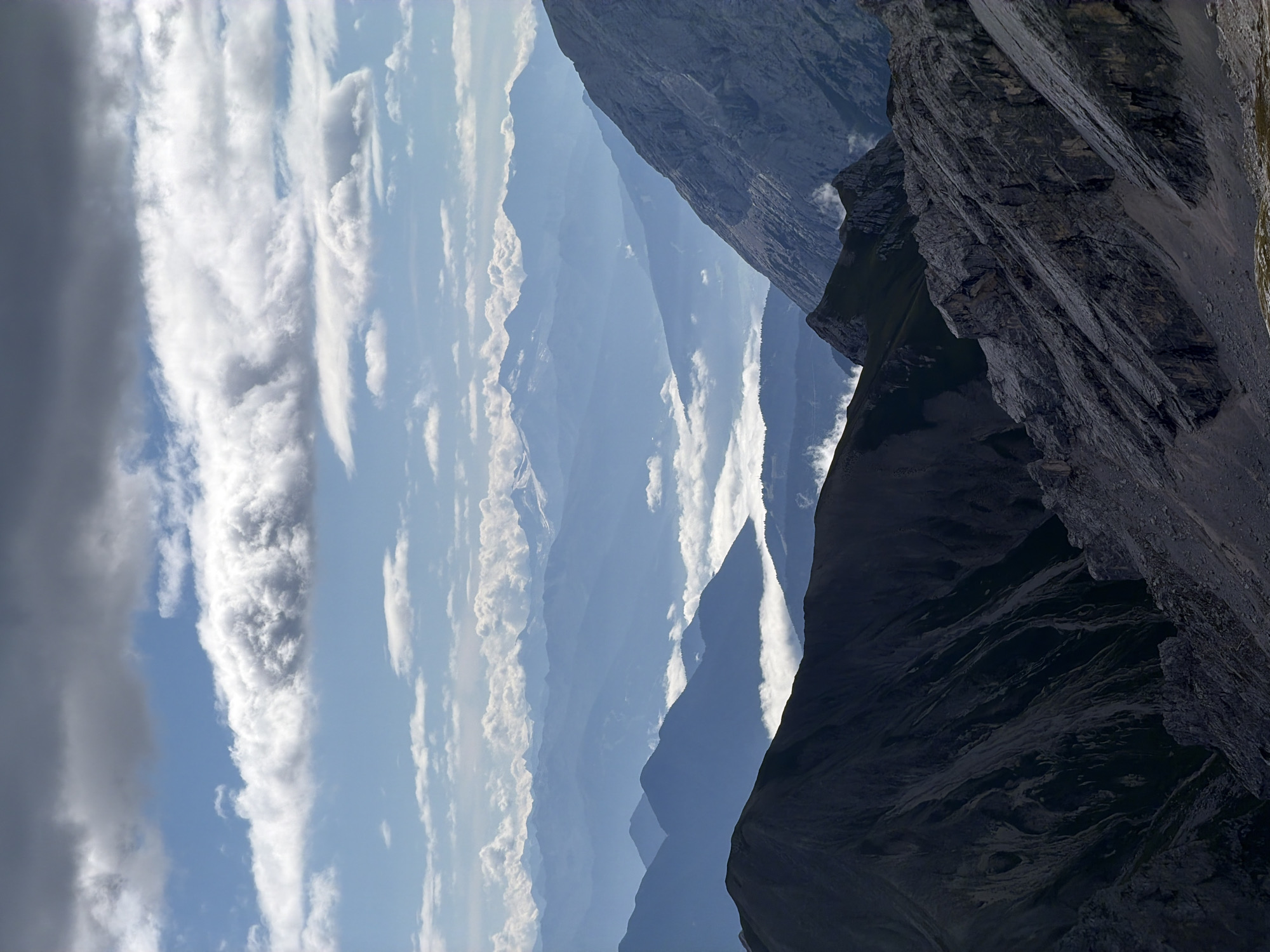 The image depicts a mountain landscape under a cloudy sky. The foreground features rugged, rocky terrain with sharp inclines and shadows. In the midground, several layers of mountains are visible, progressively fading into a hazy blue due to atmospheric perspective. The farthest mountain range is lightly dusted with snow. A layer of white clouds blankets the valleys between the midground mountains. The sky above is a mix of dark, heavy clouds and patches of bright blue sky with scattered white clouds. The scene is devoid of any people or man-made structures, suggesting a remote natural environment. While the image is a scenic mountain vista, its specific location in Garmisch-Partenkirchen, Germany cannot be determined from the visual information alone.