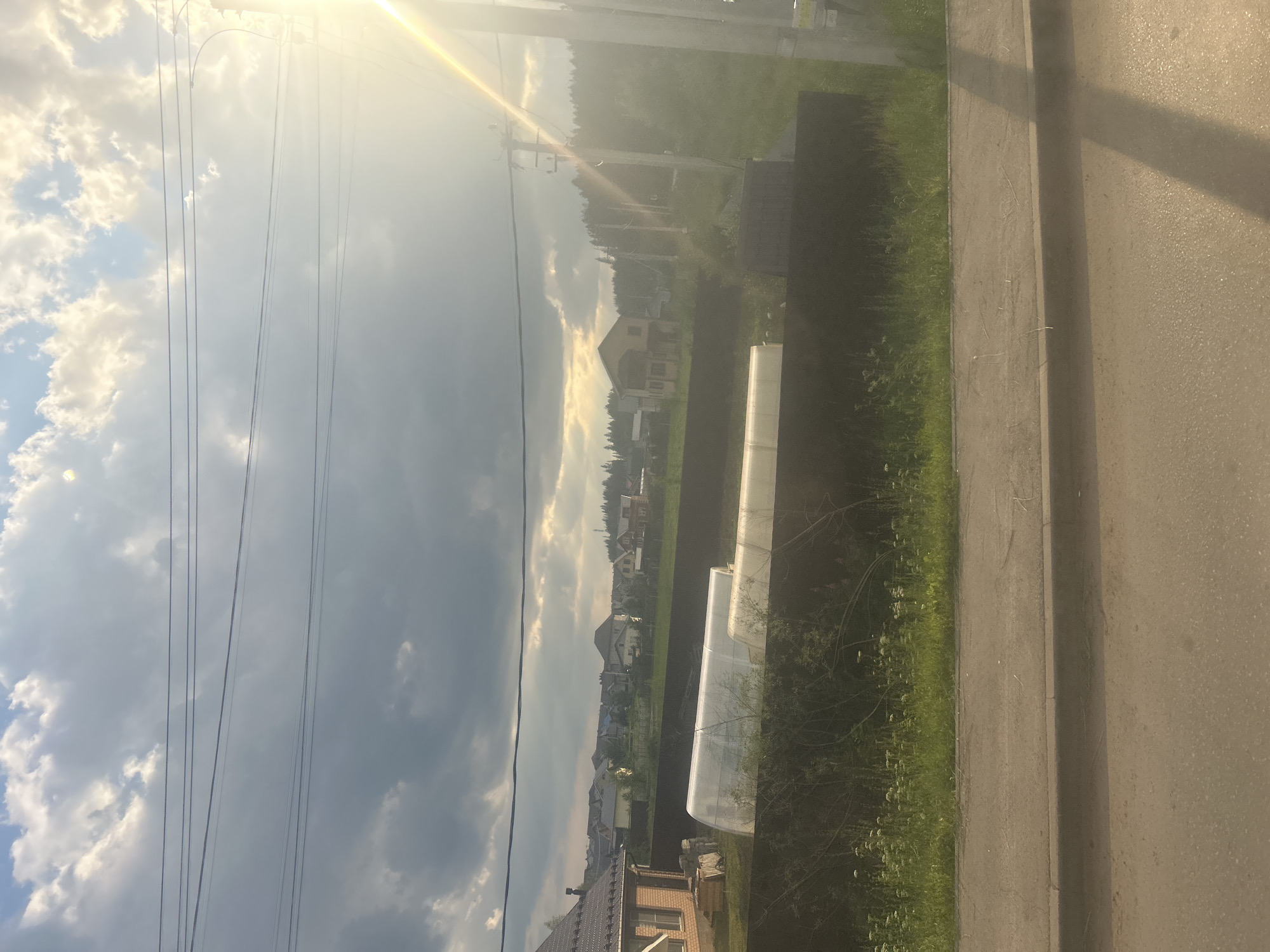 A photograph taken from a moving vehicle shows a suburban landscape in Kultaevo, Russia. In the foreground, a paved road with a curb is visible, suggesting a street or pathway. Beyond the road, a dark brown fence runs horizontally across the frame, bordering a grassy area. Two elongated, translucent structures, possibly greenhouses, are situated behind the fence, illuminated by natural light.

Further in the background, a cluster of houses with various architectural styles and roof types are spread across a gentle slope. Some houses are brick, while others appear to be painted in lighter colors. The setting sun casts a warm glow and a bright flare on the right side of the image, peeking through a partly cloudy sky. Power lines are strung across the upper portion of the frame, with utility poles also visible. The overall scene depicts a tranquil, residential area during what appears to be late afternoon or early evening.