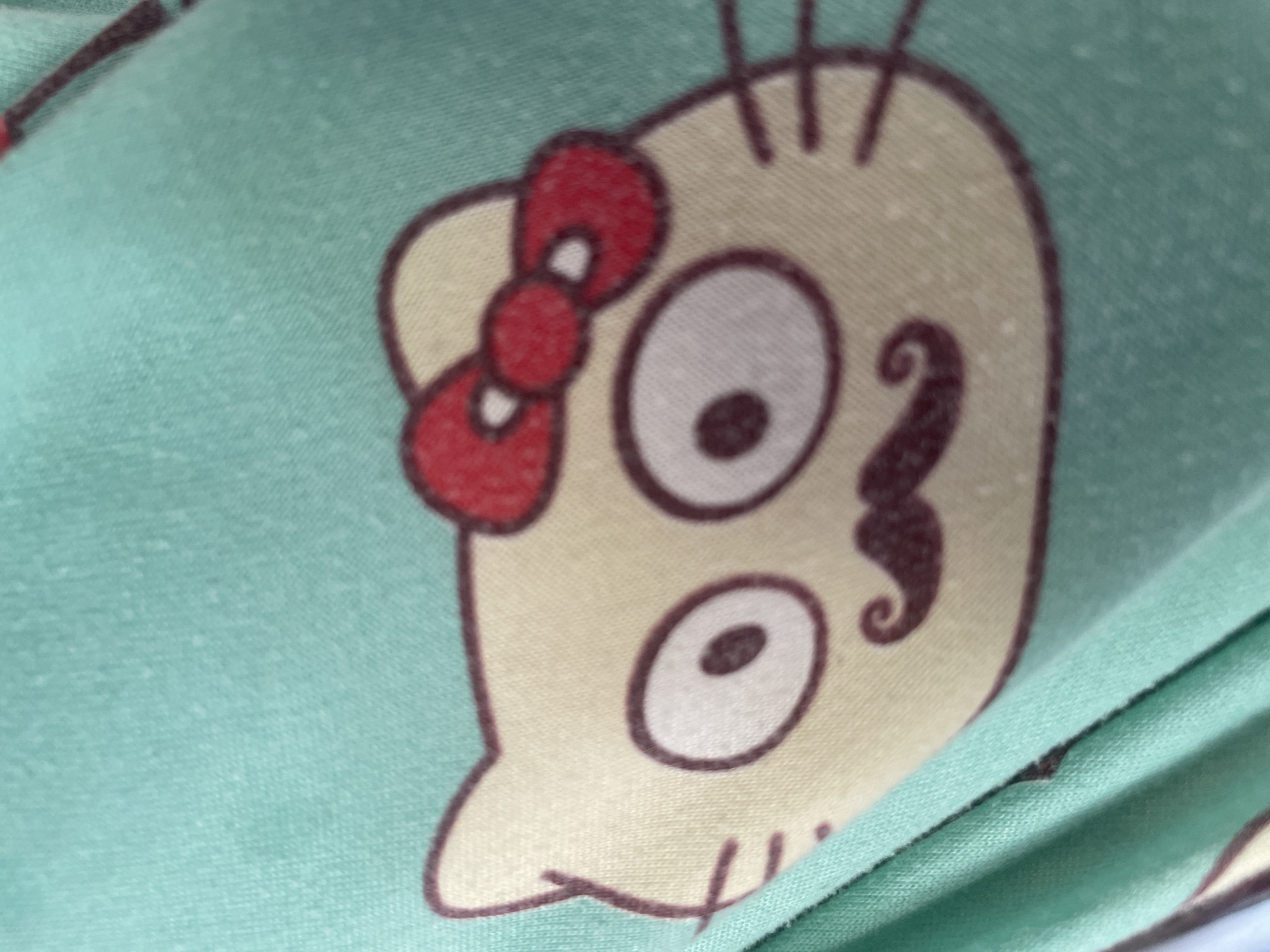The image shows a close-up view of a light green fabric with a cartoon character printed on it. The character appears to be a stylized skull with a red bow on its head, large eyes with dark pupils, and a swirl design resembling a mustache below its nose area. The fabric is folded, creating a diagonal line across the lower right portion of the image. The printing is slightly pixellated and the fabric shows some texture. There is no discernible location in Tashkent, Uzbekistan, or any other specific environmental details visible.