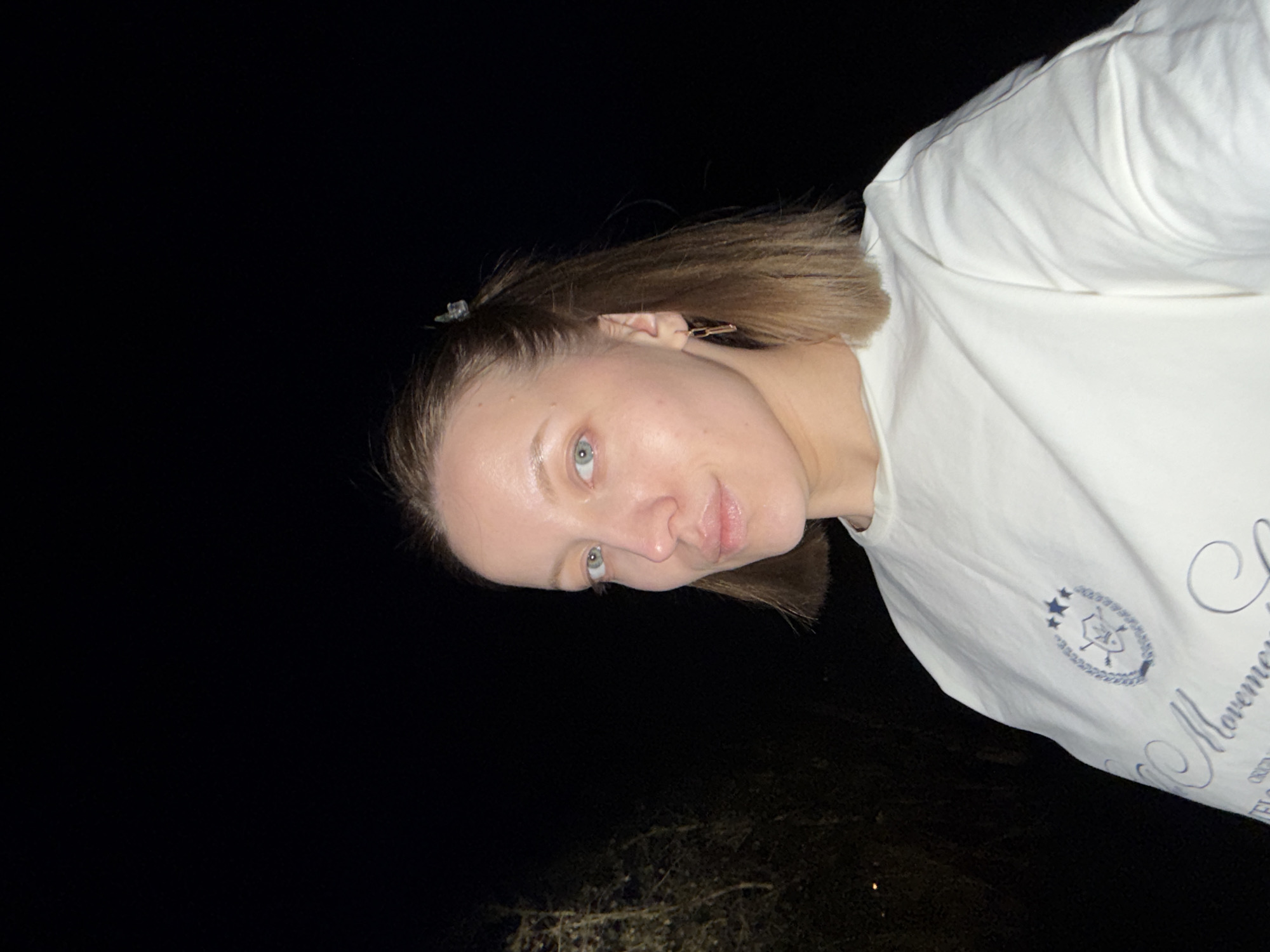 A woman with light skin and brown hair stands outdoors at night. She wears a white t-shirt with a dark logo and text. The location is potentially Reshetnikovo, Russia, but there are no visual indicators in the image to confirm this. The environment is dark, with limited visibility of surrounding objects.