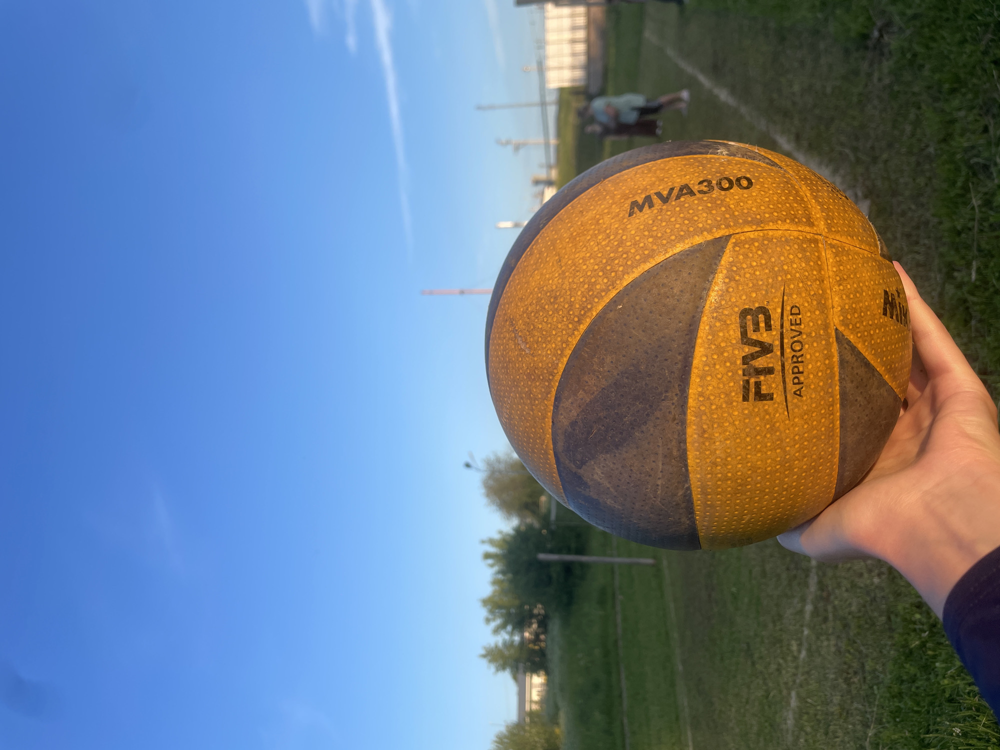 In a grassy outdoor area, a hand holds a volleyball. The volleyball is yellow and black with the text "FIVB APPROVED" and "MVA300" visible. The background shows a clear blue sky, green trees, a white building, and a few blurry figures of people standing on a court. The scene appears to be in the city of Podshivalovo, Russia, possibly on a sports field.