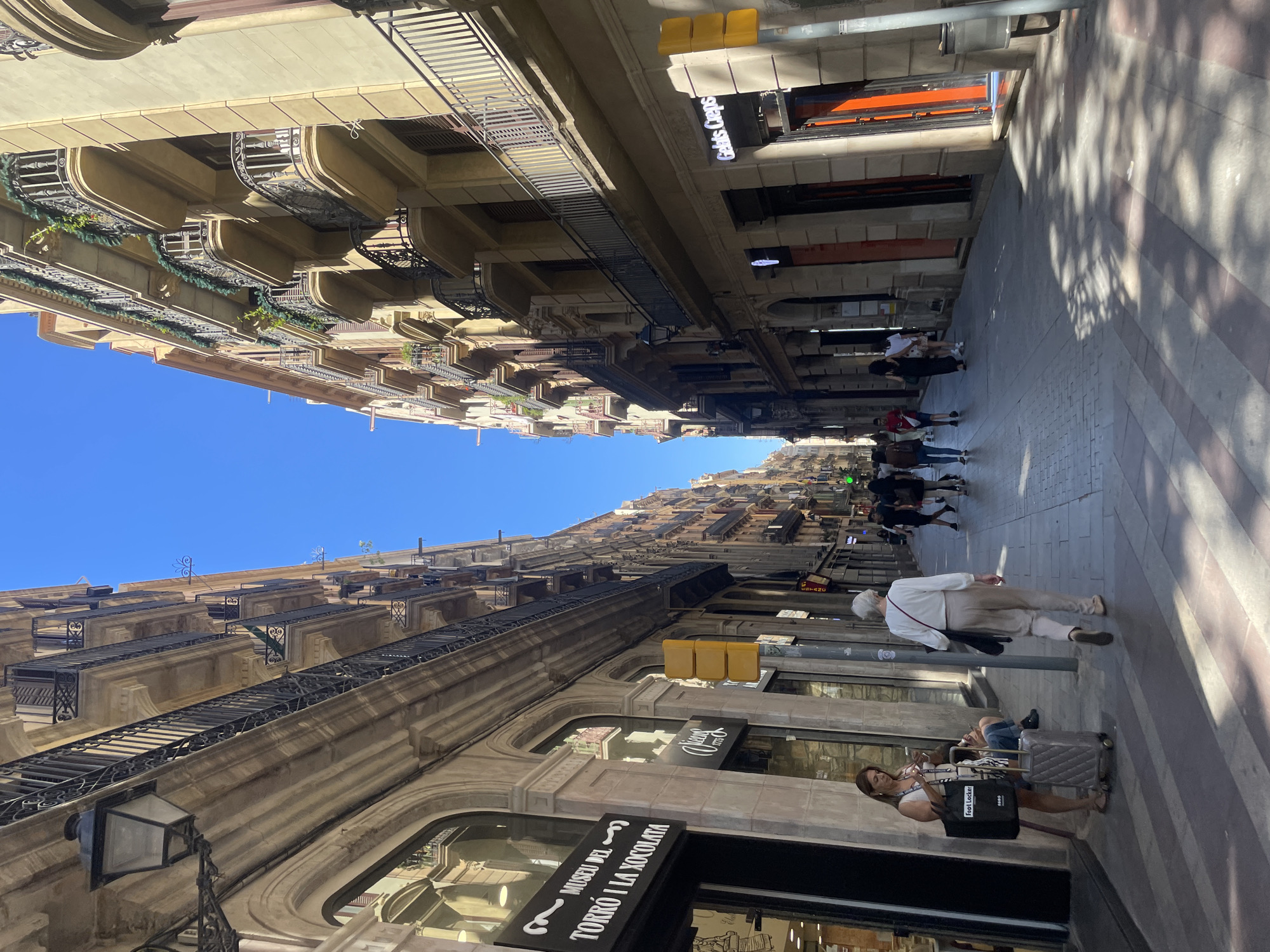 The image shows a street in Barcelona, Spain, lined with tall buildings. The buildings are characterized by multiple floors, balconies with ornate railings, and a beige to light brown color scheme. The street is paved with a striped pattern of light and dark stones. Several people are walking along the street. Some are individuals, while others appear to be in small groups. One woman is near the Museu del Torró i la Xocolata (Noucentista building) and leaning against a suitcase. Signs for shops are visible on the ground floor of the buildings, including "Gelaaati Di Marco" and "Museu del Torró i la Xocolata." The sky above is clear and blue. Street lights are present on both sides of the street.