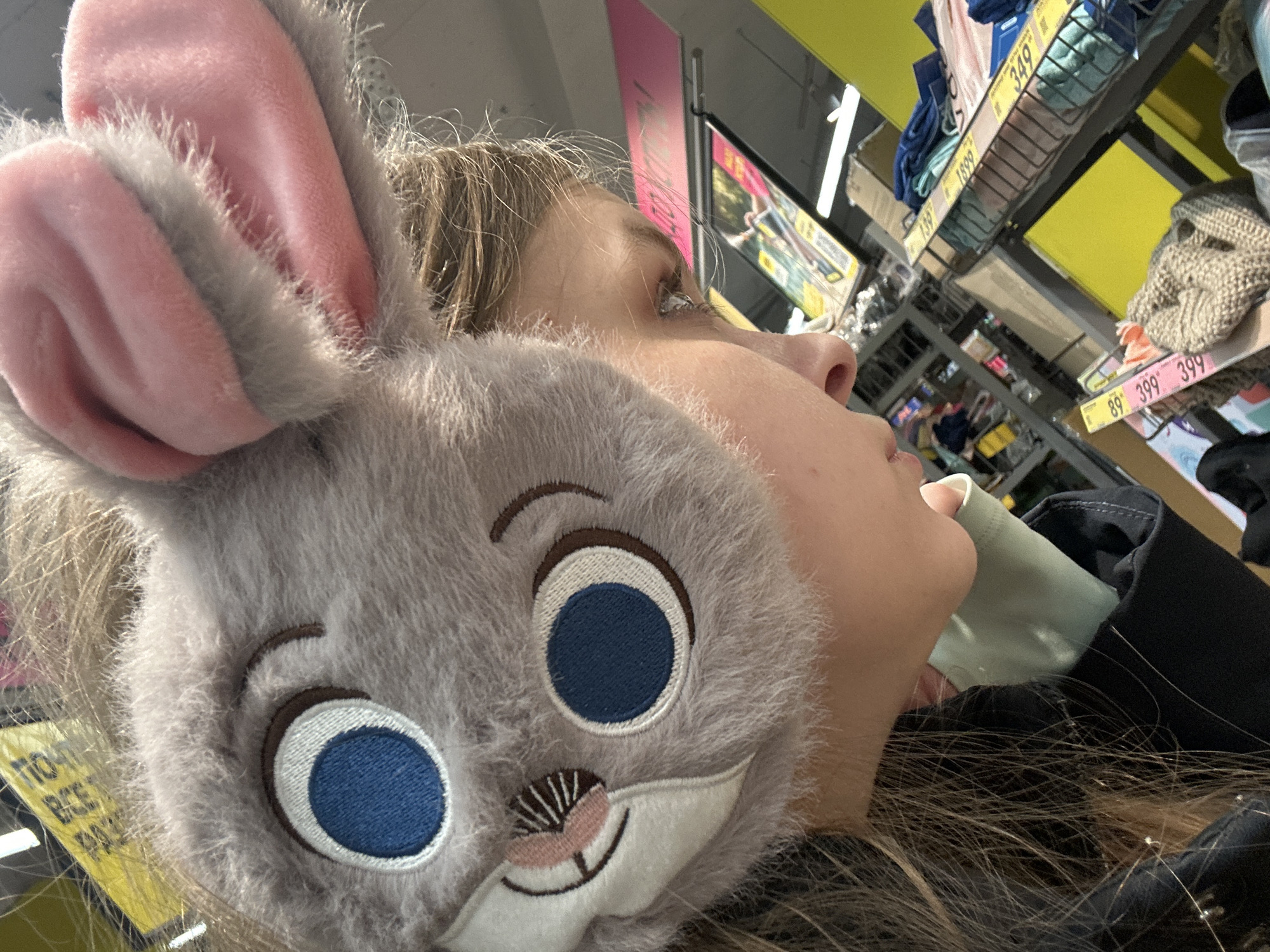 A close-up, tilted shot depicts a person wearing a plush grey bunny-ear headband with pink inner ears and a cartoon face with blue eyes and an embroidered nose and whiskers. The person is wearing a light green face mask partially obscuring their face. The background shows shelves of merchandise, including textiles, with price tags in Russian rubles. The setting appears to be a retail store, possibly in Moscow, Russia, based on the currency and the Cyrillic script on the price tags.