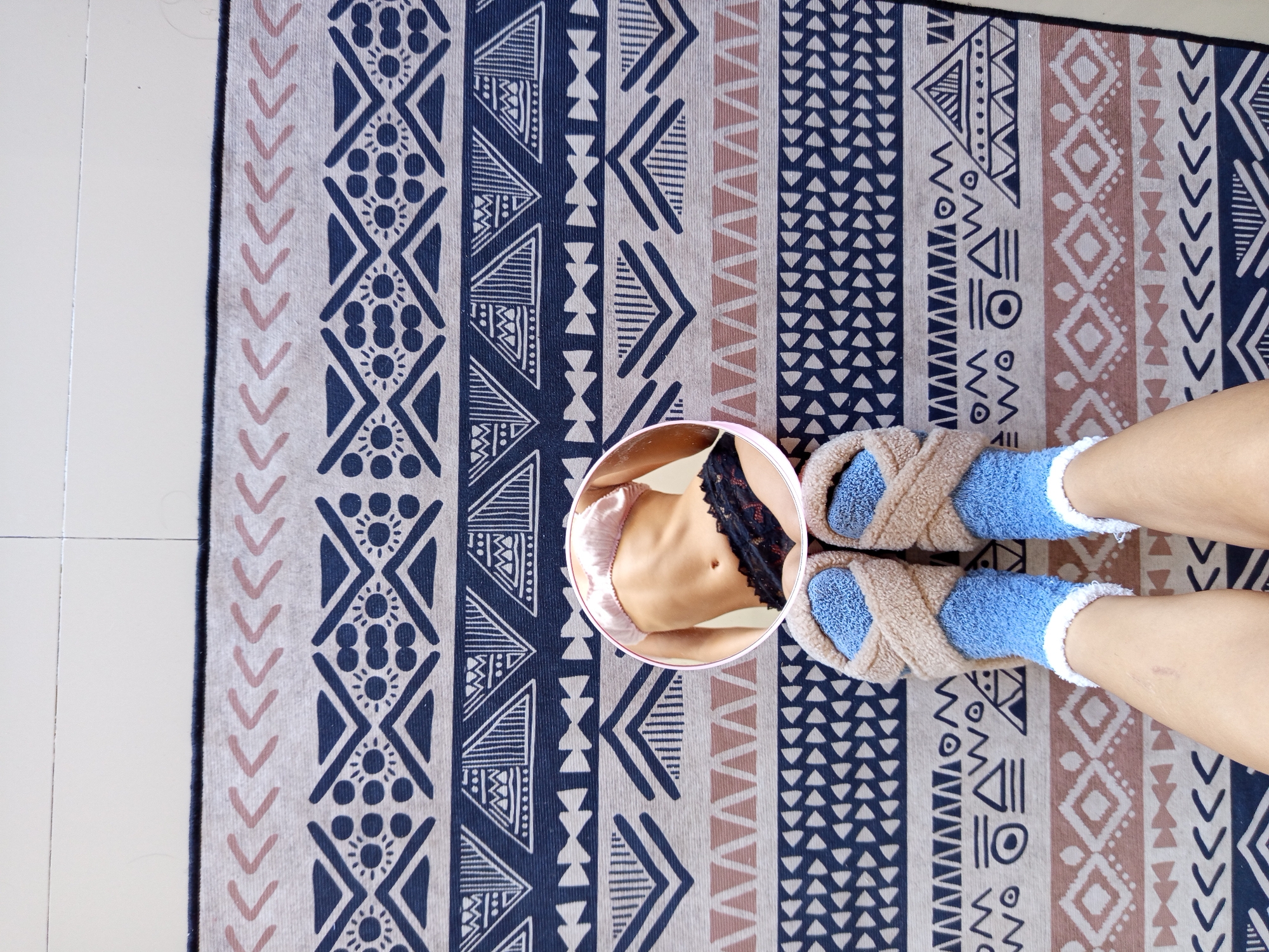 A top-down shot depicts a patterned rug with blue, brown, and white geometric shapes on a white tiled floor. Two legs wearing light blue socks and tan slippers are visible at the right. At the rug's center, a round mirror reflects a partial view of a torso wearing pink shorts and black lace lingerie. The location may be in North Kuta, Indonesia.