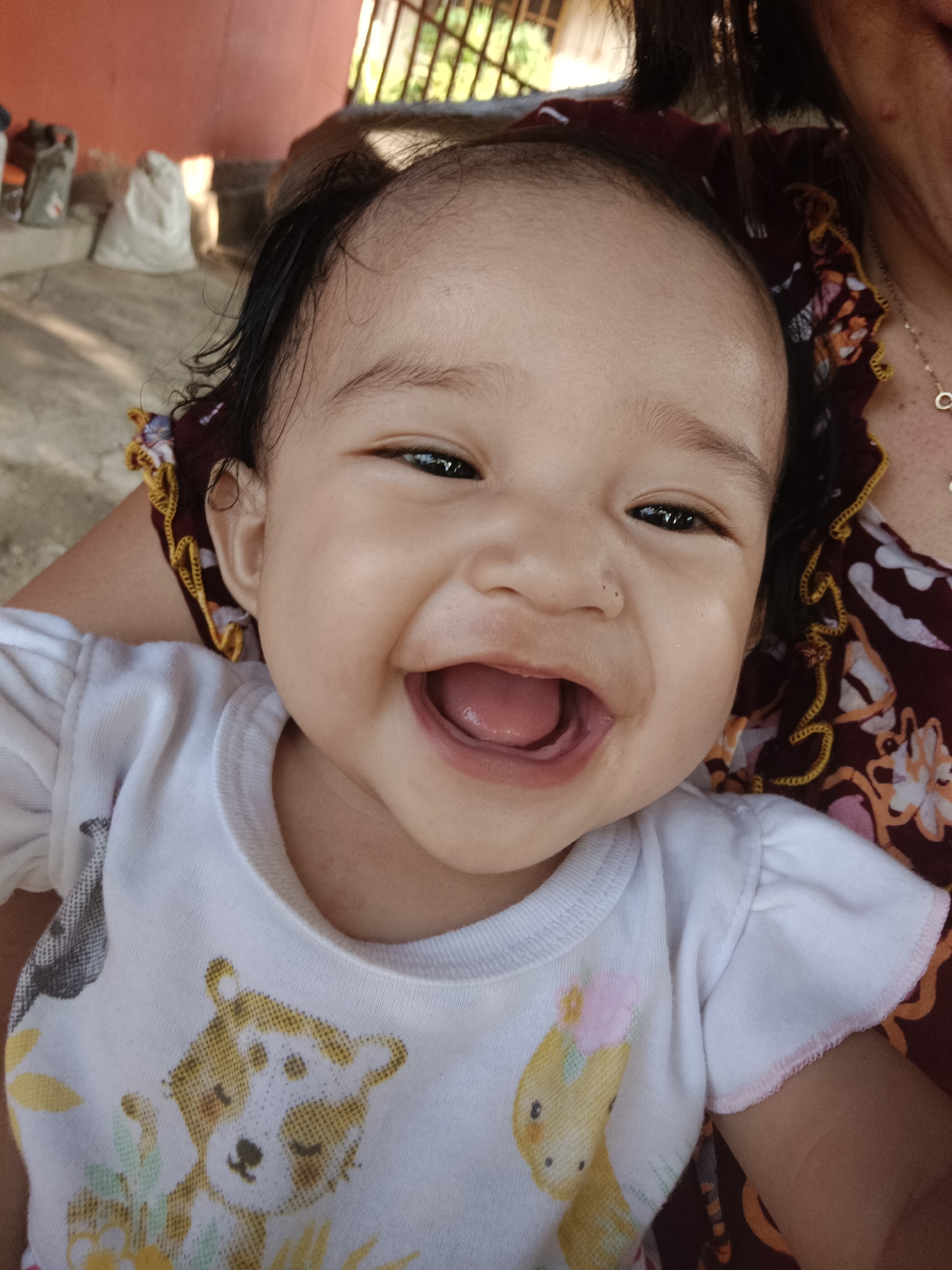 A close-up, waist-up shot shows a baby smiling widely, with its mouth open, revealing its tongue. The baby is wearing a white t-shirt with animal prints. The background is out of focus and shows a reddish-orange wall and some bags. This scene appears to be in an outdoor setting, possibly a yard or patio, in the city of Kawua, Indonesia.