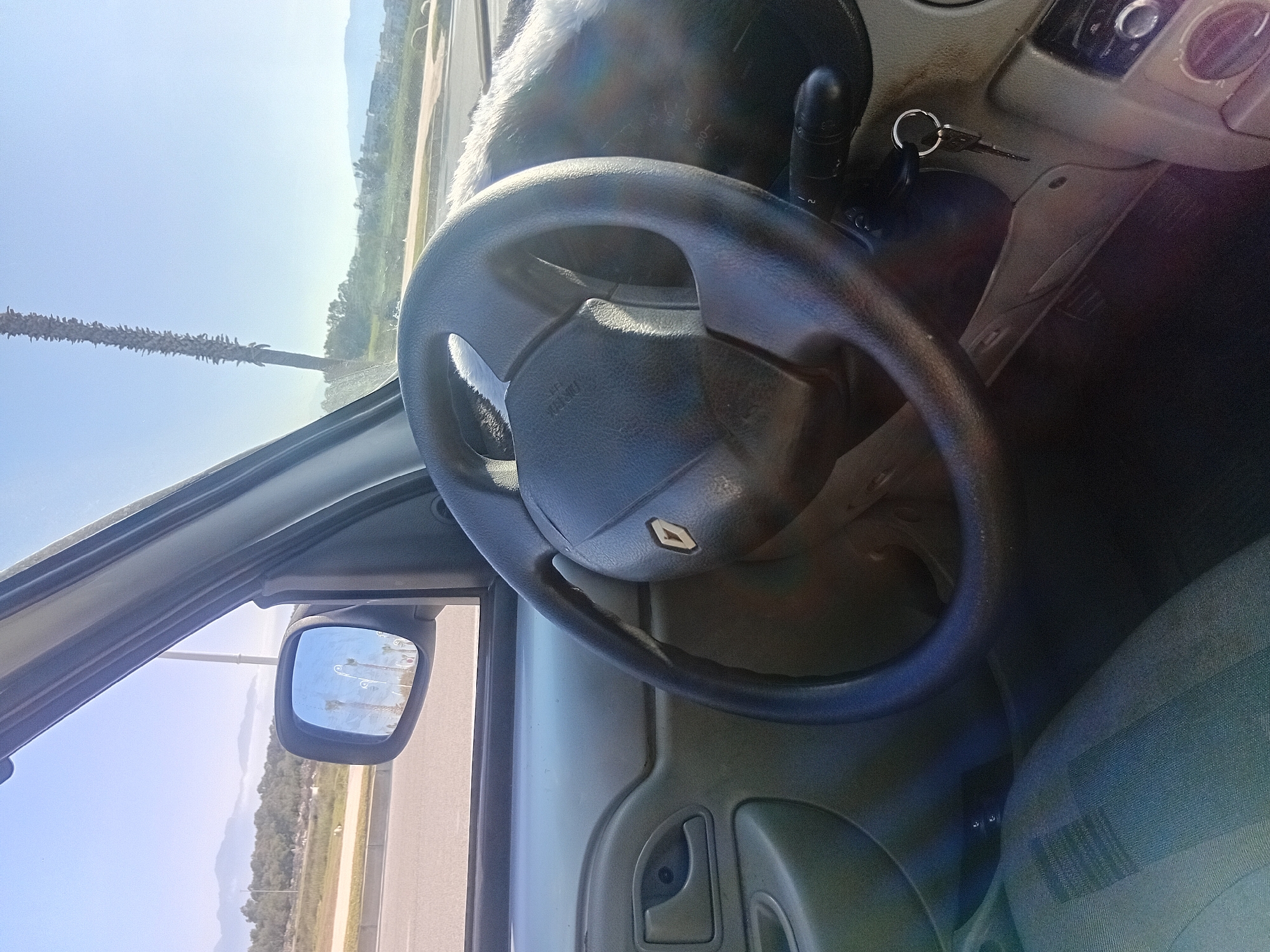 The image shows the interior of a Renault vehicle, focusing on the steering wheel with the Renault logo. The keys are in the ignition. The side mirror shows a view of a road with palm trees, suggesting a sunny day. The location is indicated as Martil, Morocco, based on visual elements consistent with the region. The image captures a static scene, likely taken from the driver's perspective while the vehicle is stationary.