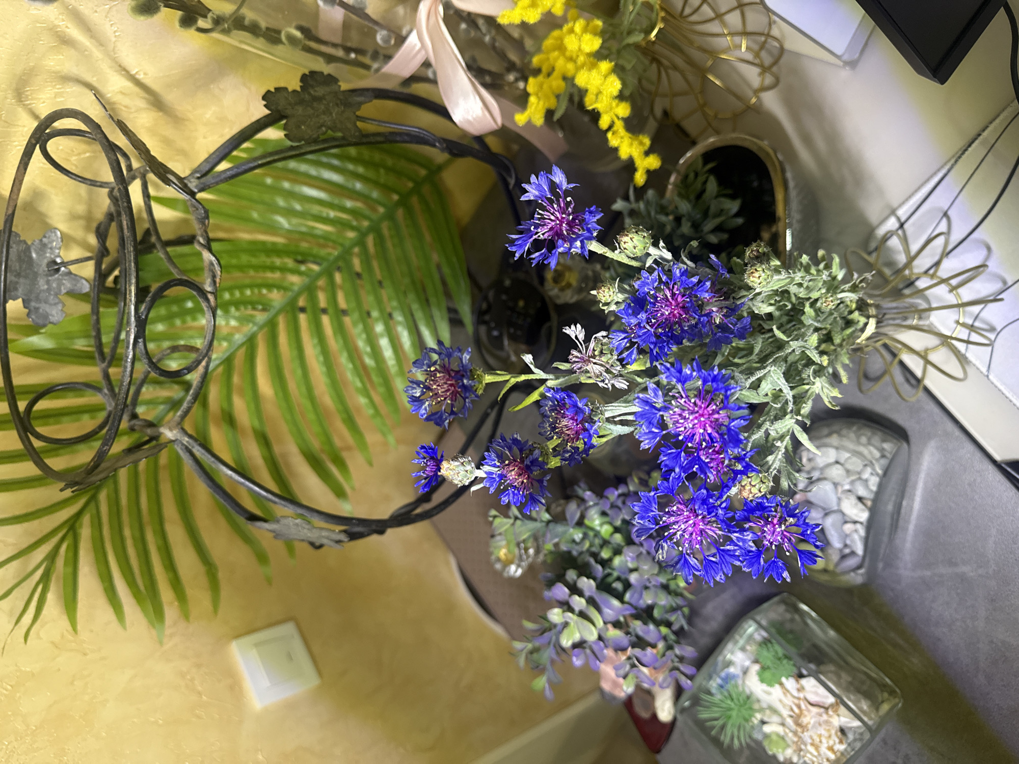 The image depicts a still life composition featuring a variety of floral elements and decorative objects. A wrought iron frame with decorative leaf accents surrounds a large, artificial palm frond. Adjacent to this are arrangements of blue cornflowers, yellow mimosa, and other greenery in several vases. The vases vary in material, including clear glass, a stone-like texture, and one with gold wire accents. The background consists of a yellow wall with a light switch. The location appears to be indoors, in a residential setting, likely in the city of Unknown, Unknown.