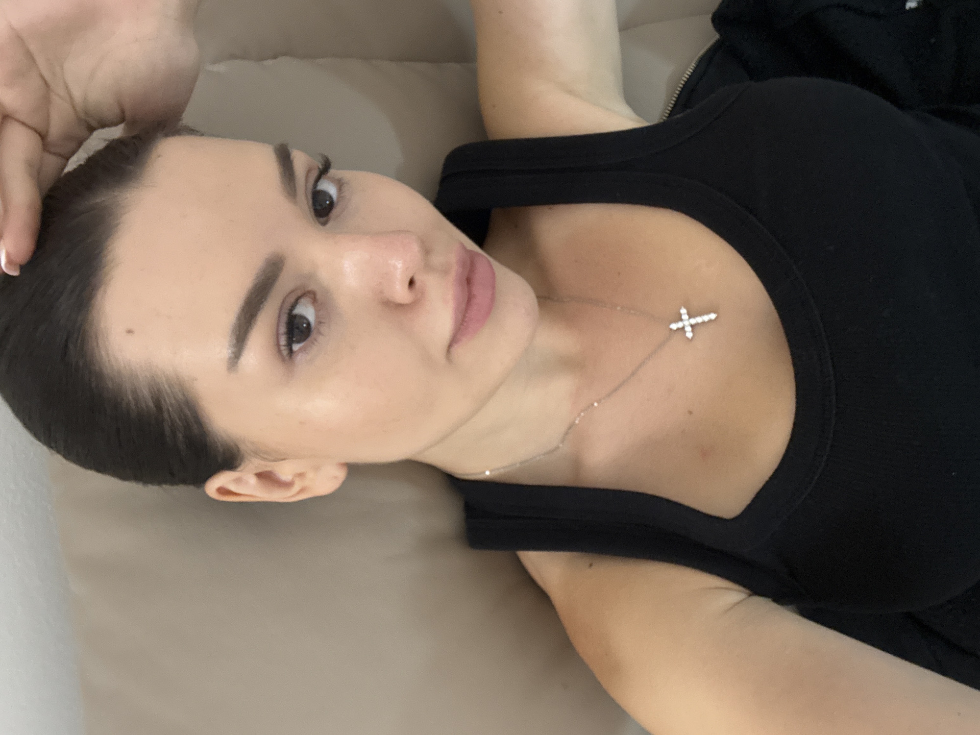 The image shows a close-up selfie of a woman. She is wearing a black tank top and a silver necklace with a cross pendant. Her hair is pulled back, and she is touching her head with her right hand. The background is a blurred neutral color, possibly a sofa or wall. The image does not provide any information about the location in Prague, Czechia.