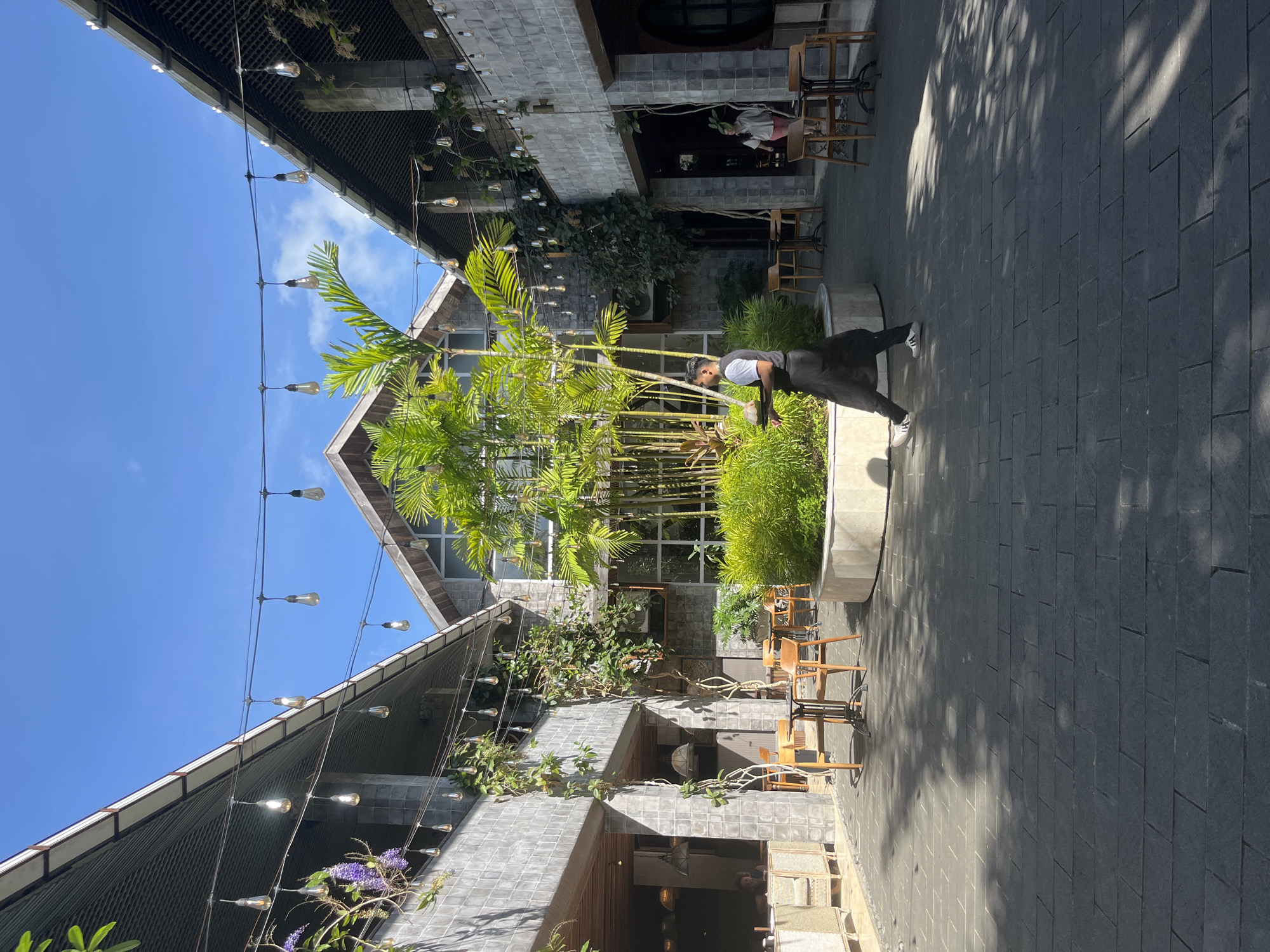 The image depicts an outdoor restaurant or café space in Petulu, Indonesia. The buildings feature gray tiled exteriors and gabled roofs, adorned with hanging light bulbs strung between the structures. Lush tropical plants, including bamboo and palm trees, grow in raised planters. A waiter in a dark uniform, a white shirt, and white tennis shoes carries a tray. Patio furniture, including wooden chairs and tables, is arranged on a dark gray stone-tiled courtyard. The sky is blue and relatively cloudless. There are patrons seated in the background of the restaurant.