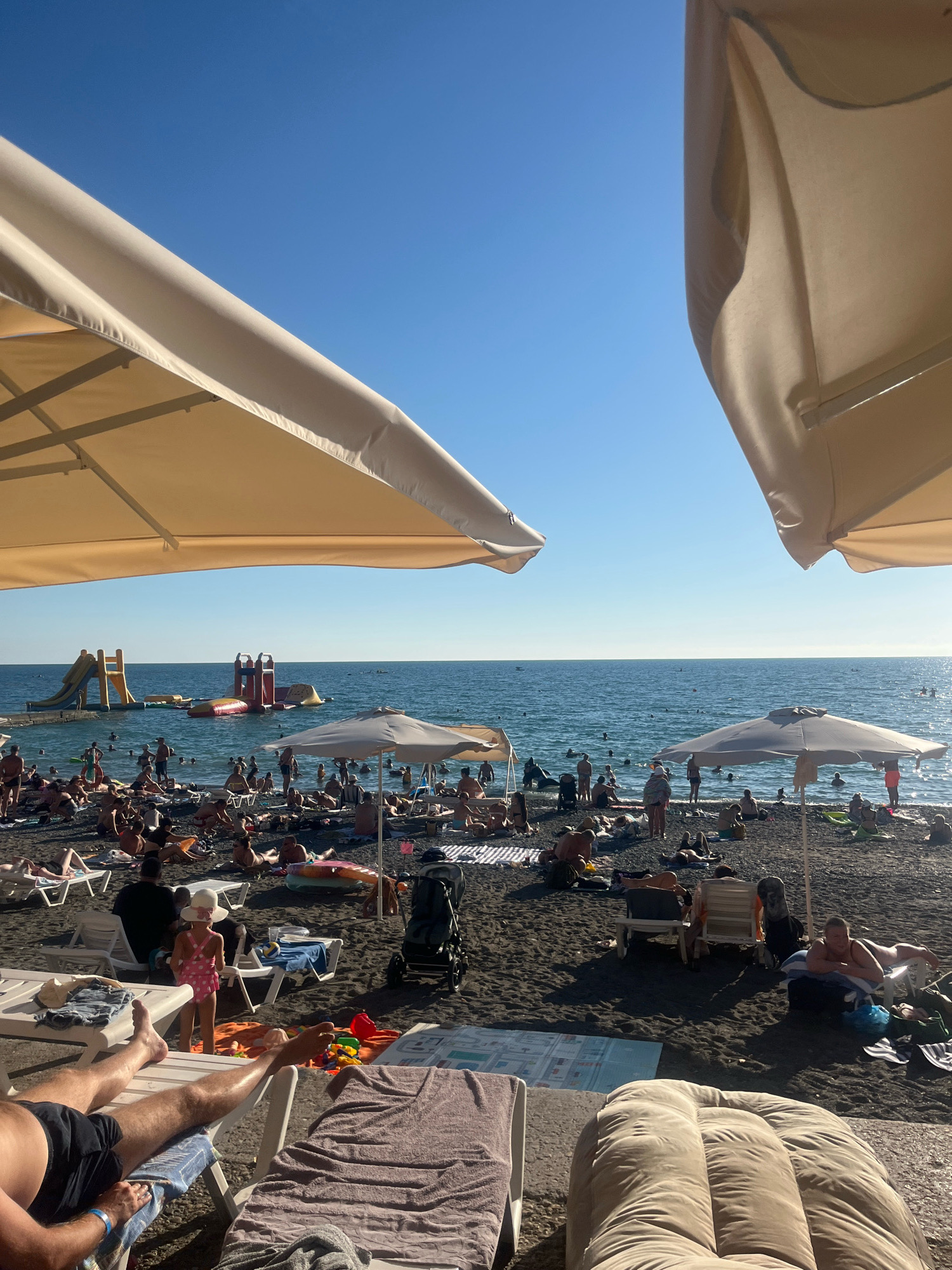 This is a wide-angle, eye-level shot of a crowded black pebble beach on a sunny day. The sky is clear blue and takes up about one-third of the frame. Two large, light-colored umbrellas dominate the foreground, partially obscuring the view of the beach and the sea. The beach is populated with numerous people sunbathing on lounge chairs, towels, and directly on the pebbles. Many people are in the sea, some swimming and others near inflatable water slides and attractions.
In the middle ground, to the left, a yellow and blue inflatable water slide is set up in the water, with people visible climbing on it and sliding down. To the right of that, a red and white inflatable water attraction is also visible. A few people are walking along the shoreline, and others are sitting in lounge chairs under smaller umbrellas scattered across the beach. A child in a pink polka-dot swimsuit and a white sun hat stands near the center of the image, facing away from the camera. Several strollers are present on the beach.
The sea is calm with small waves, and the sunlight glints off the water. The overall atmosphere is one of a busy summer day at a popular beach resort. This scene is consistent with beaches found in Sochi, Russia, known for its pebble beaches and recreational water activities.