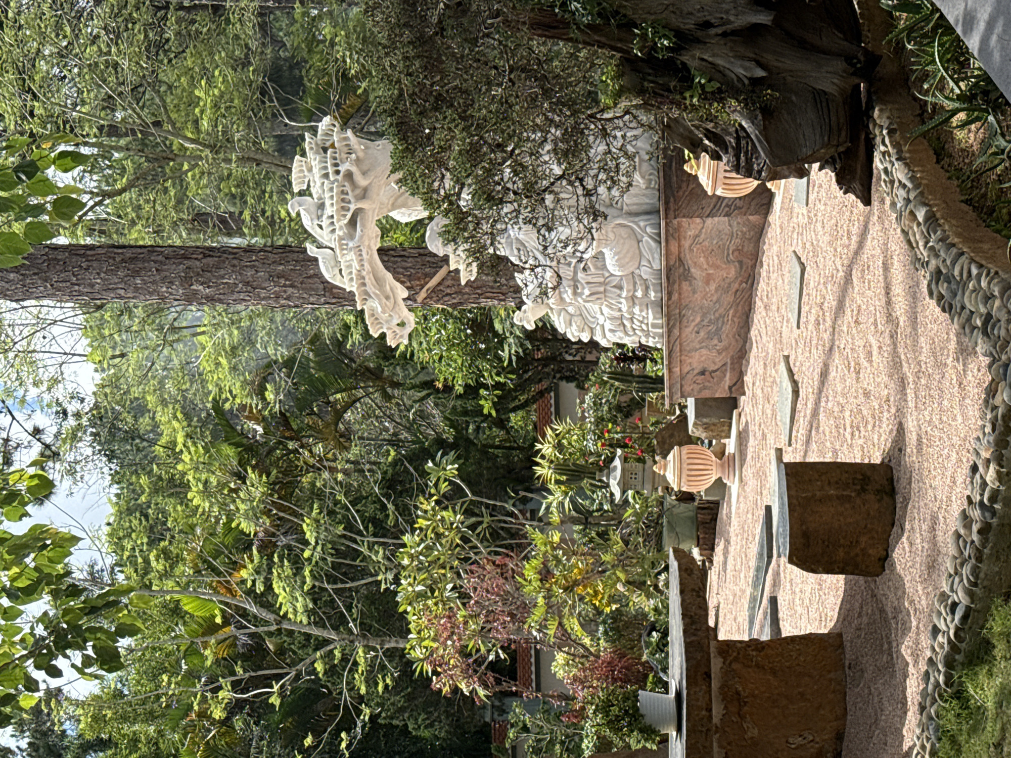 This image depicts an outdoor Zen garden with a stone table and stool set on a bed of pink gravel. The garden features lush greenery, including trees and various plants, and a large white marble sculpture of a dragon or phoenix, accompanied by birds, positioned behind a large tree trunk. To the right of the sculpture, there is a weathered, gnarled tree trunk. Two decorative urns and a stone lantern are also visible. The scene suggests a tranquil, meditative atmosphere. This garden is located at the Thien Vuong Zen Monastery in Da Lat, Vietnam.