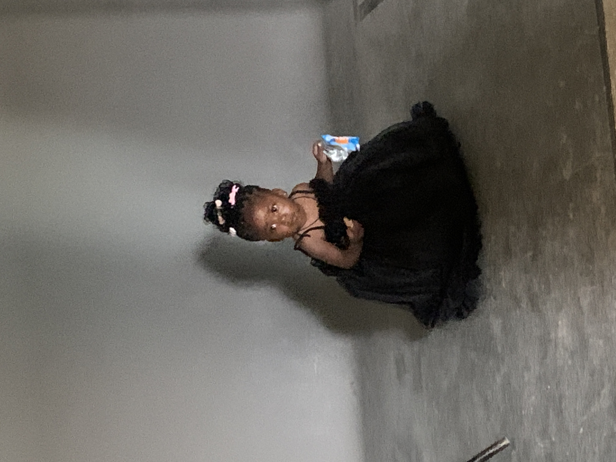 A young girl in a black dress stands in an interior space with gray walls and flooring. She is holding a water bottle. The location is Gura Top, Nigeria.