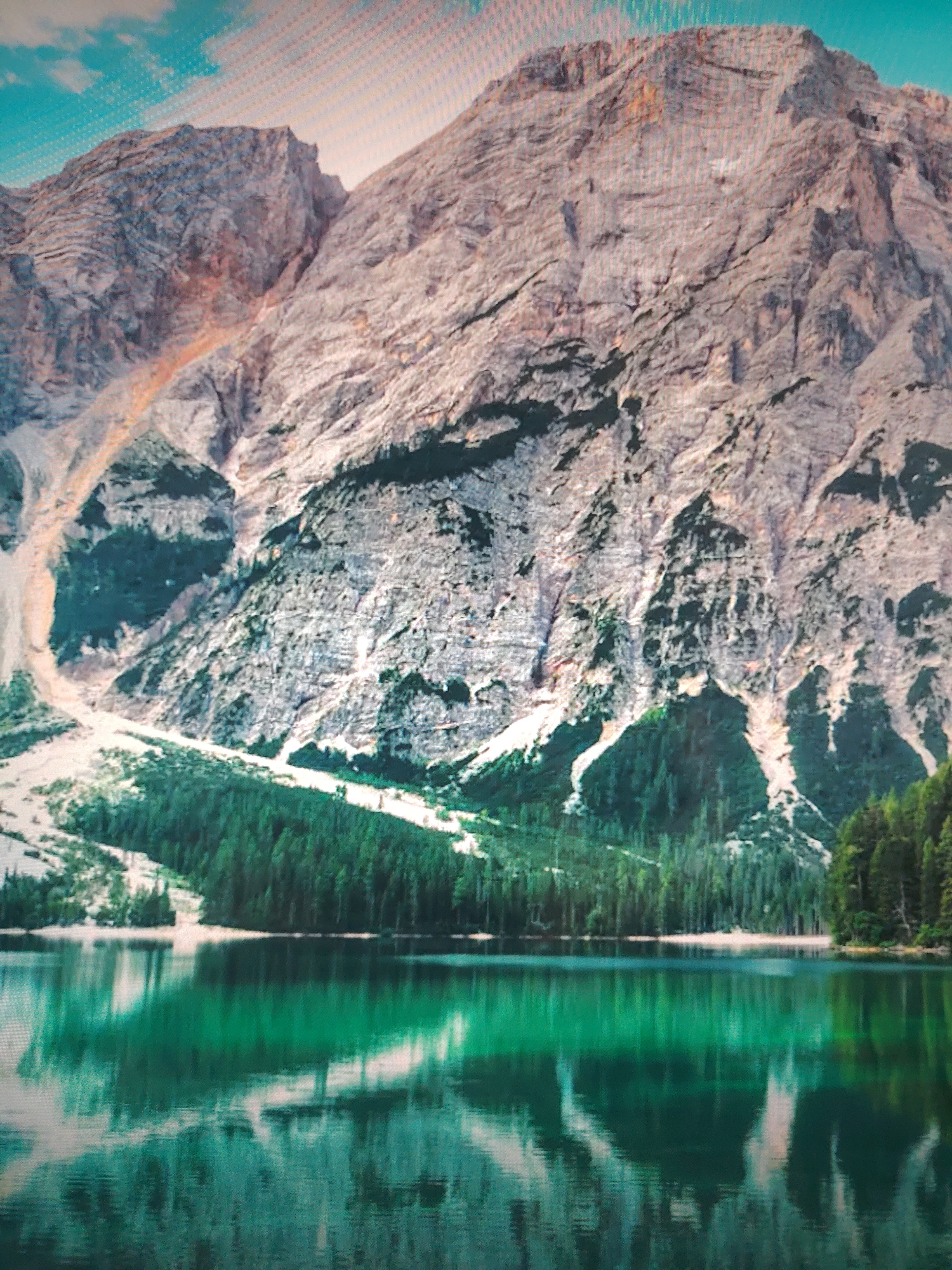 The image shows a mountain range reflected in a body of water. The mountains are rocky and sparsely vegetated, with some green patches. A forest lines the edge of the water, and the water itself is a deep green color. The sky is visible in the upper left corner, with a hint of blue. The location is unknown.