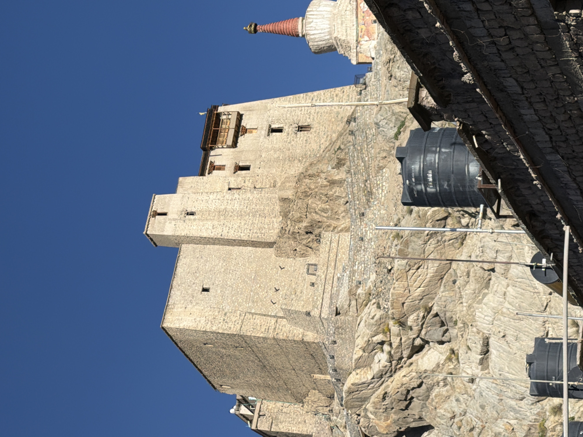 This is a low-angle, vertical outdoor shot of the Leh Palace, a historic royal palace in Leh, Ladakh, India. The palace is a multi-story stone structure built on a steep hillside, with a prominent central tower. A Buddhist stupa with a red spire is visible to the right of the palace. The sky is clear and blue. In the foreground, there are some black water tanks and parts of a stone building with a textured roof. The scene is brightly lit by the sun, suggesting it is daytime. There are no people visible in the image.