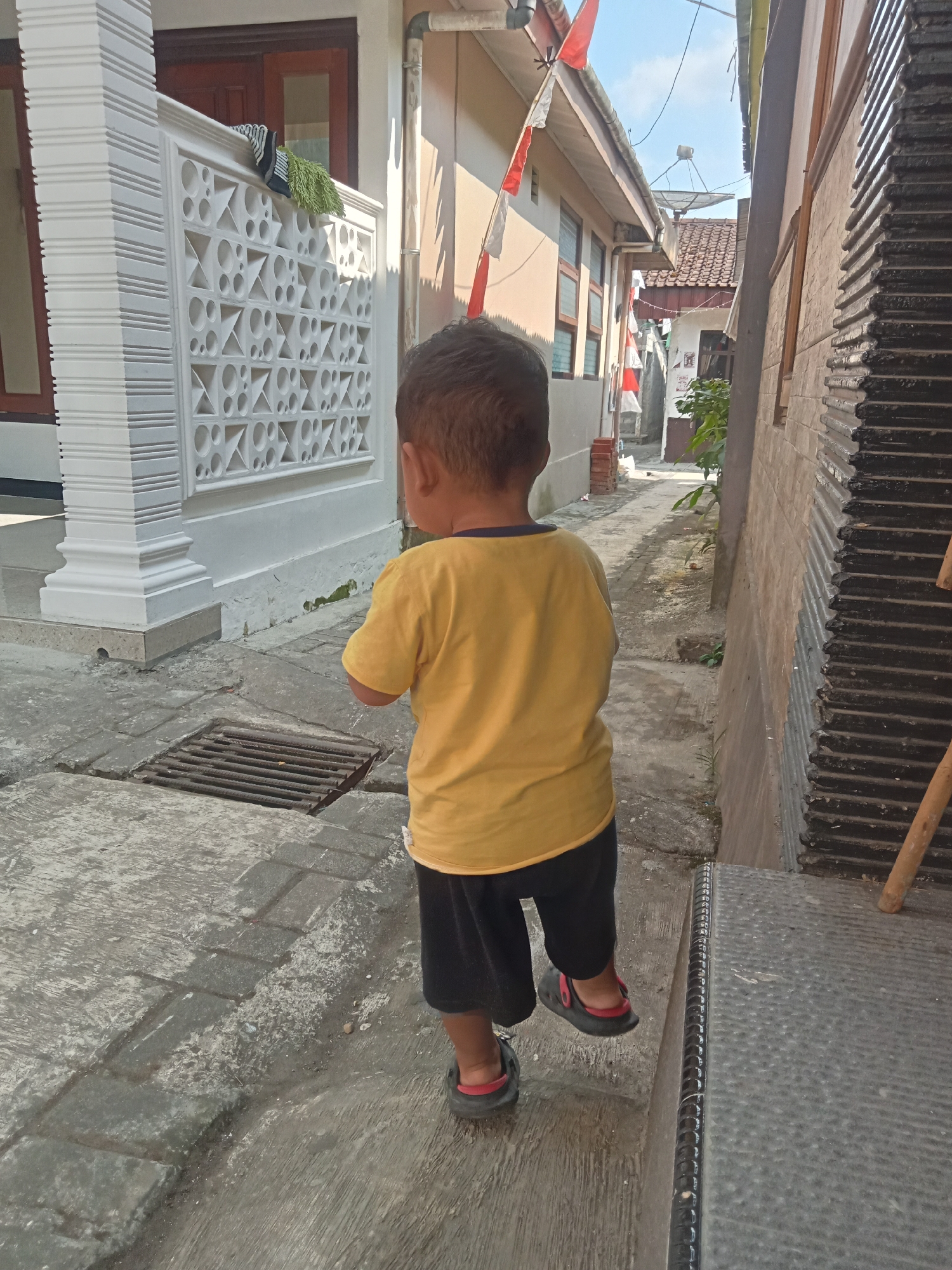 A young child, viewed from behind, walks down a narrow outdoor pathway between two buildings. The child wears a yellow t-shirt and black shorts with red-accented slip-on shoes. To the left of the pathway is a white decorative wall with a patterned design, a white pillar, and a metal grate set into the paved ground. The buildings appear to be single-story structures with tiled roofs and some red and white flags are visible on the left building. The pathway is paved with interlocking gray stones and concrete, with some areas showing wear and debris. To the right, a dark, ribbed metal structure lines the side of the pathway. The sky is visible above, indicating an outdoor, daytime setting. The location is identified as Pancurwening, Indonesia.