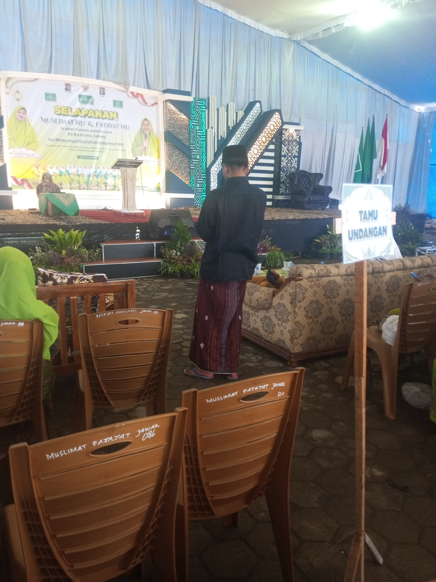 This image depicts an outdoor event likely held in a semi-public area in Klaseman, Indonesia, possibly a village hall or community center, given the presence of tents and makeshift stages. The scene is dominated by a stage set up with a backdrop banner featuring text in Indonesian, indicating a Muslim gathering called "SELAPANAN MUSLIMAT NU & FATAYAT NU SE-AKABANG MOJOTENGAH DI RANTING JAWAN." The banner also includes images of women and text related to a religious ceremony.
In the foreground, several plastic chairs are visible, many with handwritten labels on their backs, such as "MUSLIMAT PATAYAT JAWAR 086" and "JAWAH 90." These likely denote seating arrangements or group affiliations. A person wearing a black shirt and a sarong stands facing away from the camera, looking towards the stage. Another person, wearing a green hijab, is seated on the left side of the image.
Behind the chairs, a patterned sofa is positioned, suggesting a seating area for distinguished guests. Flags, including green ones and one with red and white stripes (likely the Indonesian flag), are displayed to the right of the stage, alongside a sign that reads "TAMU UNDANGAN" (Guest Invitation). The ground is paved with interlocking stones. The overall environment suggests a moderately organized event, possibly a celebration or a religious gathering, with a mix of formal elements like the stage and banner, and informal touches like the plastic chairs. The lighting appears to be a mix of natural light and artificial lighting from the overhead tent.