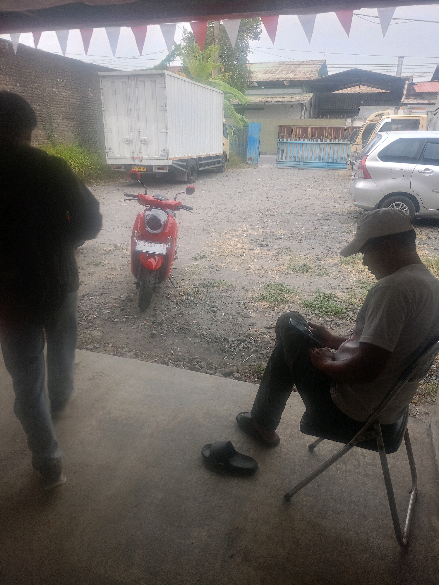 A red scooter is parked in a dusty outdoor lot. To the left, a person wearing dark clothing stands with their back to the camera. In the center-right, a person wearing a light-colored shirt and a cap is seated on a folding chair, looking at a phone. The background shows a white box truck, other vehicles, and buildings. The scene appears to be in Cirebon, Indonesia, possibly a parking area or roadside.