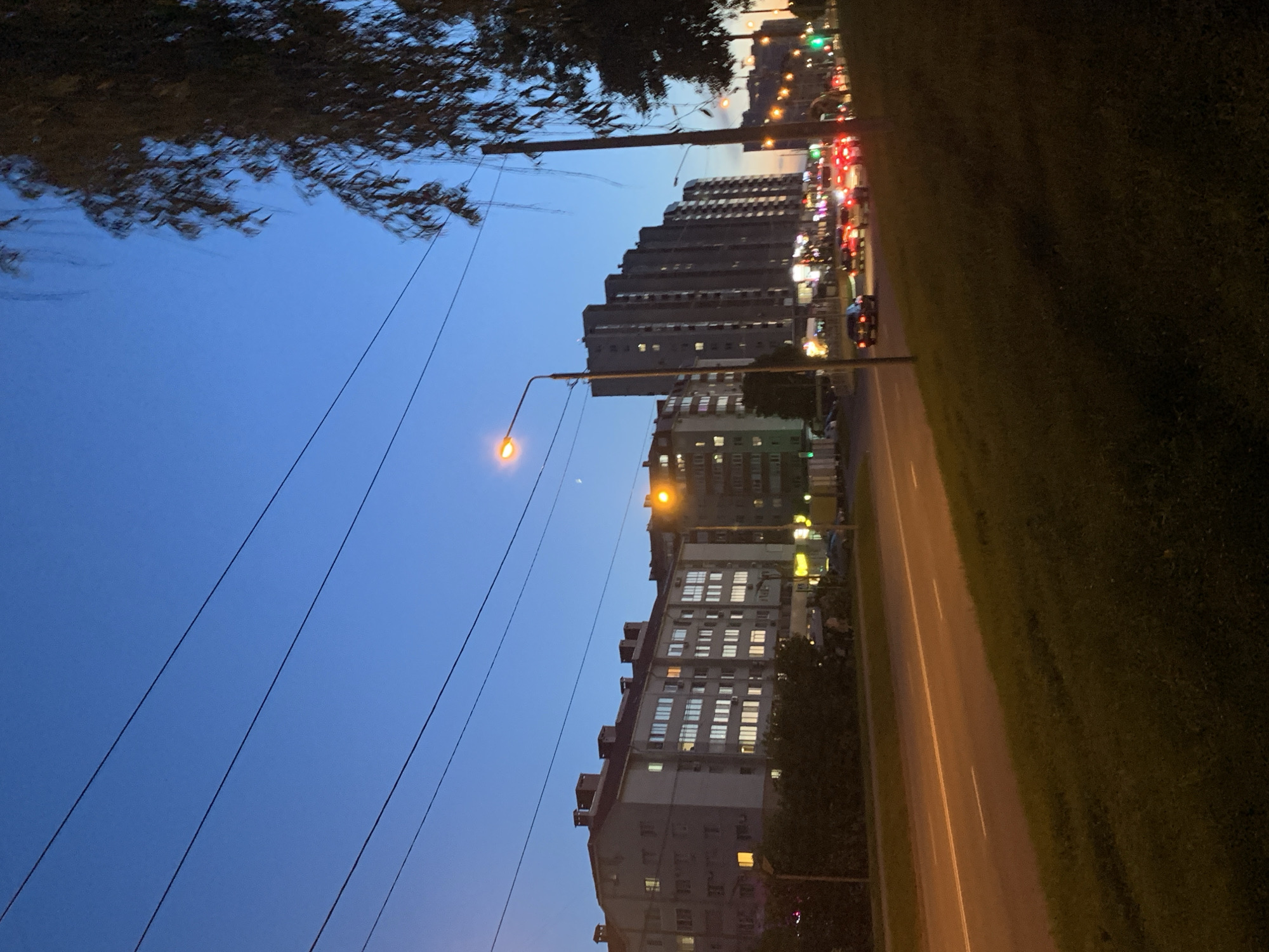 The image captures an urban street scene in Stavropol, Russia, during dusk or early nightfall. The sky, a deep twilight blue, dominates the upper left, adorned with several diagonal power lines. A faint, thin crescent moon is subtly visible against the blue sky, situated above the buildings.
On the right side of the frame, a row of multi-story apartment buildings stretches into the distance. Numerous windows in these residential structures are illuminated, suggesting occupancy. Two prominent streetlights cast a warm orange glow, one in the mid-left foreground and another further down the street.
Running along the bottom of the image is a paved road with visible white lane markings. The road appears busy, with several cars showing distinct red taillights, indicating traffic. In the background, beyond the immediate buildings and traffic, more city lights and structures are visible, extending into the evening haze. The right side of the road features a grassy verge, and dark tree foliage partially frames the top left corner of the scene. No discernible text is present in the image.