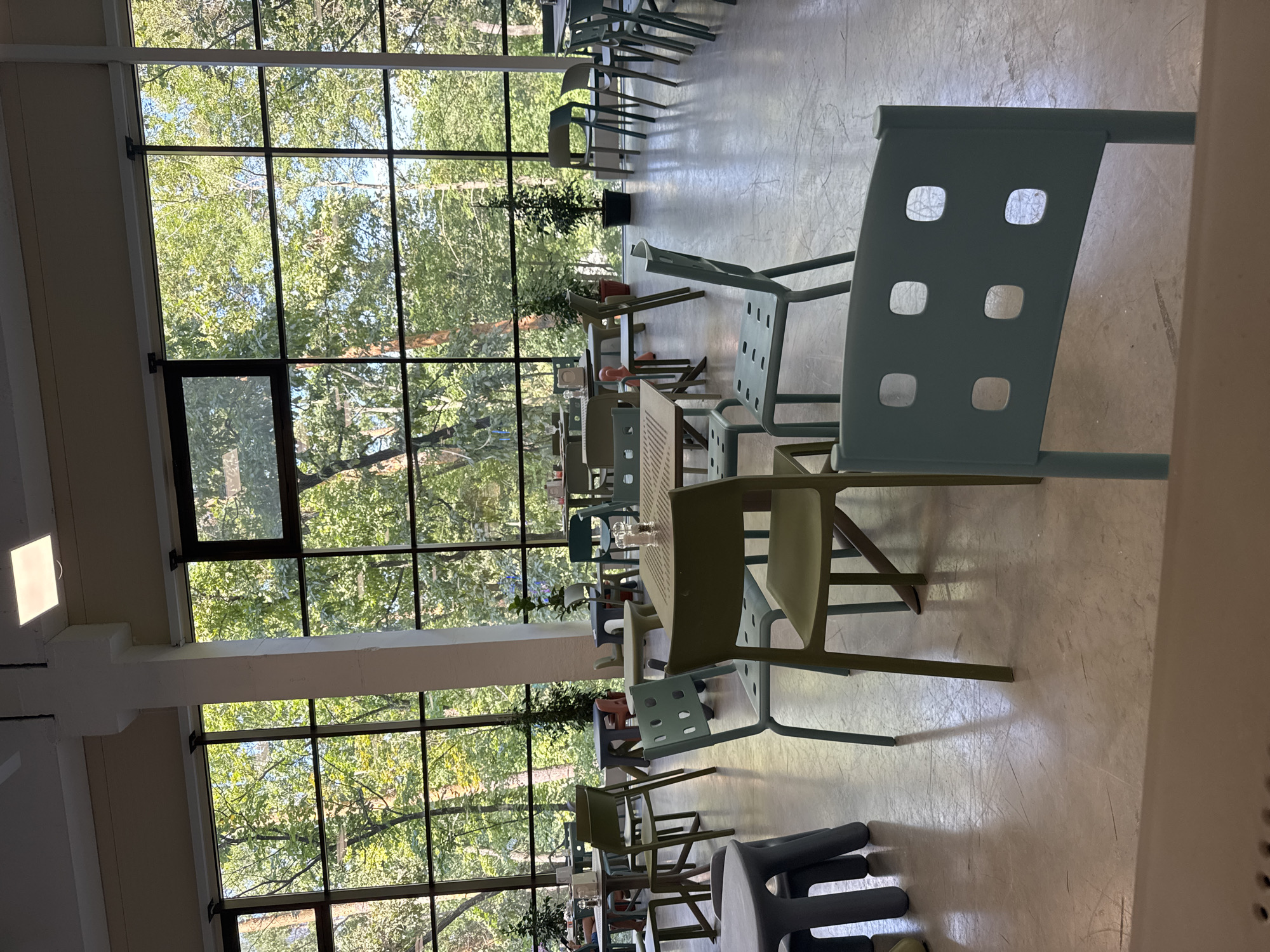 The image depicts the interior of a modern, brightly lit dining area with large windows offering a view of lush green trees. The room is furnished with numerous tables and chairs, predominantly in shades of light blue, green, and beige, creating a contemporary and minimalist aesthetic. The tables are of various shapes and sizes, some round, some rectangular, and some appear to be modular. The chairs are molded plastic, some with simple backrests and others with distinctive square cutouts. Several small, round salt and pepper shakers are visible on some of the tables. The floor is a light-colored, smooth surface, reflecting the light from the windows and overhead lighting. The presence of artificial plants suggests an indoor setting designed to bring the outdoors in. There are no people visible in the image, contributing to a sense of quietude and emptiness. The overall impression is of a clean, spacious, and somewhat sterile environment, likely a cafeteria or a modern restaurant, designed to accommodate a moderate number of patrons. The exterior view through the windows shows dense foliage, indicating that the location is surrounded by nature. No specific elements in the image definitively tie it to Orlivshchyna, Ukraine, or any particular city.