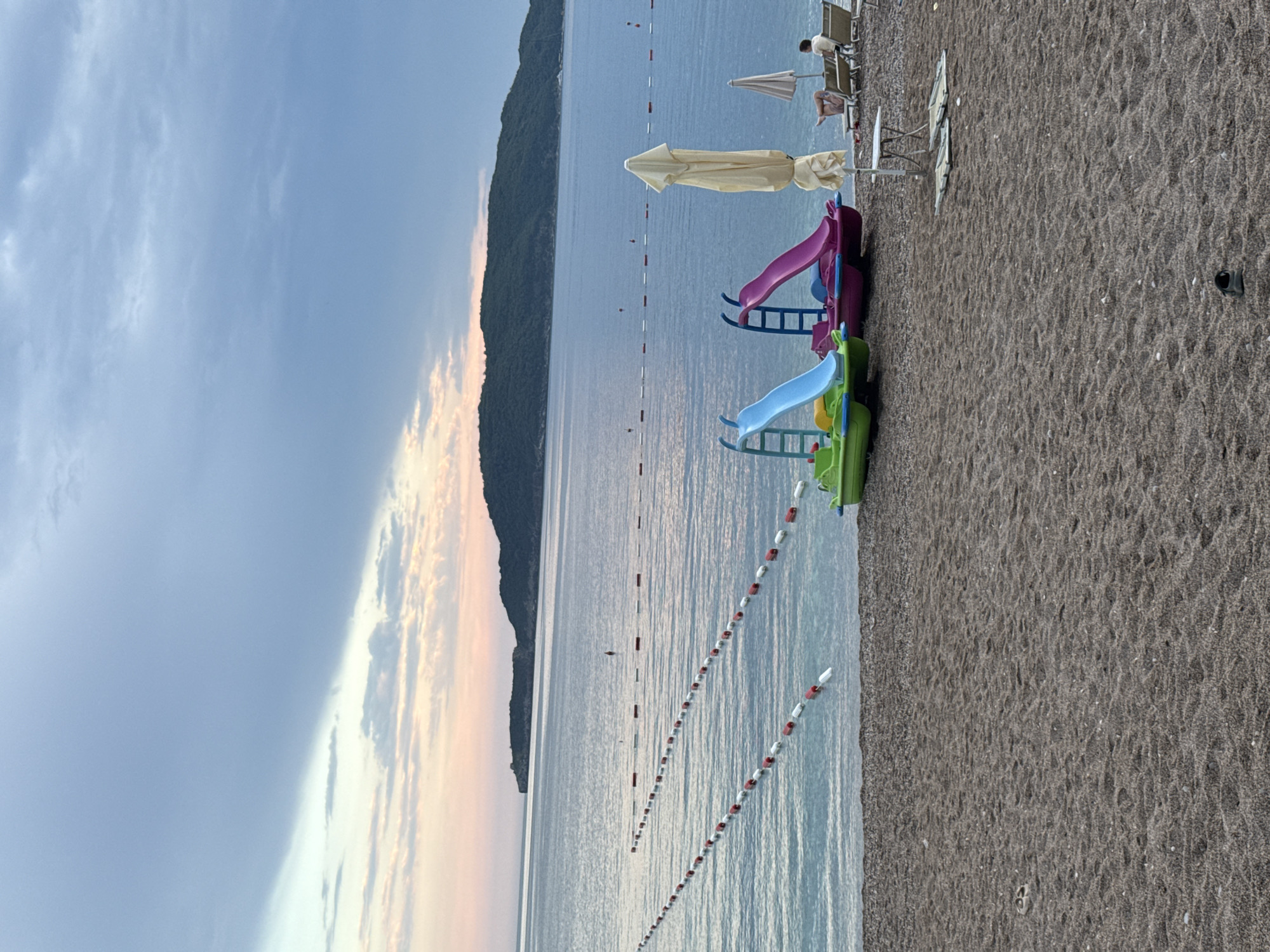 This image depicts a beach scene at Kamenovo, Montenegro, likely during the late afternoon or early evening due to the soft, warm light illuminating the sky and water. The sky is a gentle gradient of blue, with streaks of pale orange and pink clouds visible near the horizon, suggesting a sunset. The sea is calm and reflective, with ripples gently lapping against the shore.
The foreground is dominated by a dark, pebbly beach. Scattered on the beach are several beach chairs, some occupied by figures, and a small table. A few beach umbrellas are also visible, one of which is partially deployed. Closer to the water's edge, there are two colorful plastic slides, one blue and one purple, likely intended for children's play. A yellow umbrella is folded and placed behind them.
A white buoys and red marker line separates the swimming area from the open sea. In the distance, a dark, tree-covered mountain range forms the backdrop, meeting the sea on the left side of the image. The overall mood is peaceful and serene, characteristic of a relaxing day at the beach as the day winds down. No text is visible in the image.