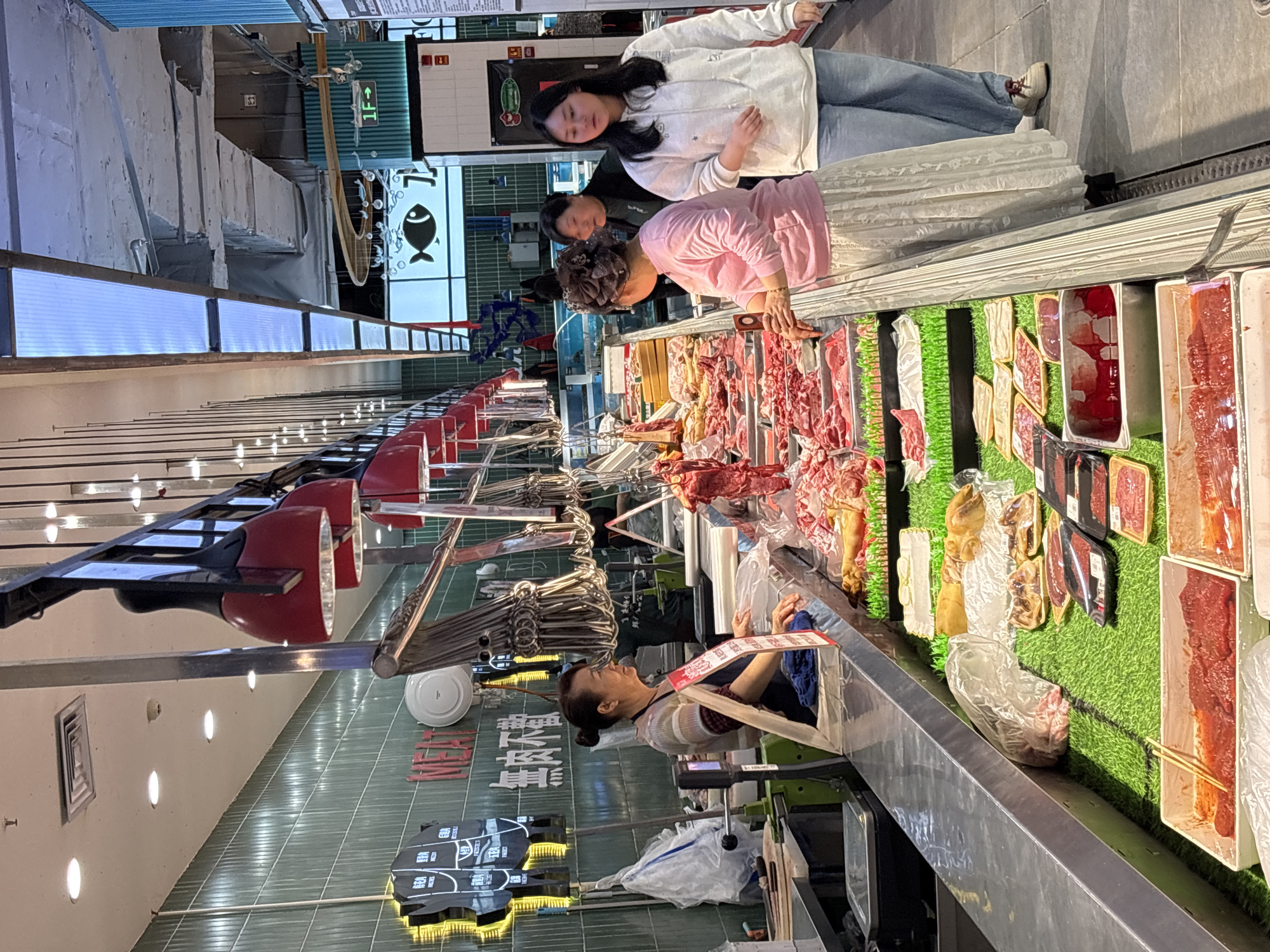 The image shows an indoor butcher shop in Jiangjin, China. A female butcher stands behind a counter filled with various cuts of meat arranged on green artificial turf. Three female customers are looking at the meat selection. The lighting is bright, and the shop appears clean and well-stocked. Overhead, red lamps illuminate the display, and hanging meat hooks are visible. The wall behind the counter is tiled in green and features Chinese characters. A sign with a fish illustration hangs near the ceiling.