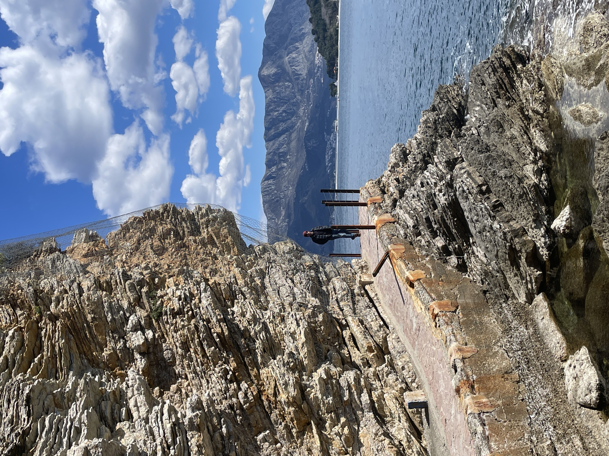 The image depicts a coastal scene along a pedestrian path in Budva, Montenegro. A weathered, stratified rock cliff, partially covered by a dark grey safety mesh, occupies the left side of the frame. A narrow, reddish-brown paved walkway, supported by a concrete structure exhibiting rust stains and erosion, runs along the base of the cliff, immediately adjacent to the sea. A low barrier composed of dark metal posts and horizontal rails lines the seaward edge of the walkway.
One individual stands on the walkway, positioned towards the right side of the frame, facing away from the camera and towards the open sea. The person is wearing a dark jacket, blue jeans, dark sunglasses, and a red beanie, with their hands clasped behind their back.
The calm, blue-green sea extends to the horizon on the right. In the distance, across the bay, prominent grey-blue mountains rise under a bright blue sky dotted with scattered white cumulus clouds. A coastal settlement with white buildings and a strip of beach is visible at the base of the distant mountains. The foreground includes rocky outcroppings and small pools of water along the base of the cliff, where the sea meets the land.