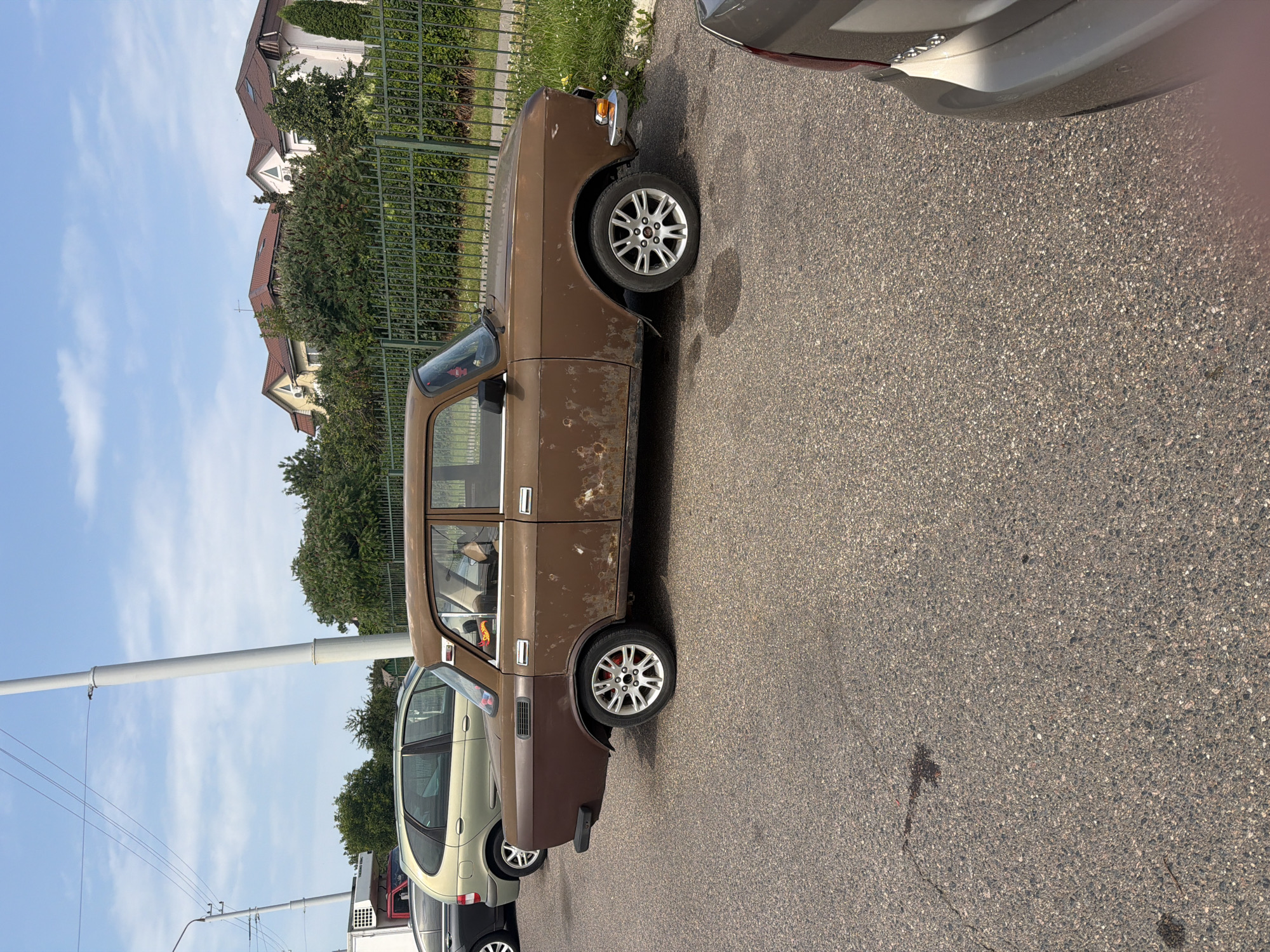 In Minsk, Belarus, on a paved street, a rusty brown vintage car is parked. The car has four doors and is seen from the side profile. It has silver multi-spoke rims with red brake calipers. The car's paint is visibly deteriorating with rust spots and peeling. Behind the car, a green metal fence runs along a strip of grass and trees. In the background, several houses with red-tiled roofs are visible, set against a clear blue sky with scattered white clouds. Other vehicles are parked nearby.