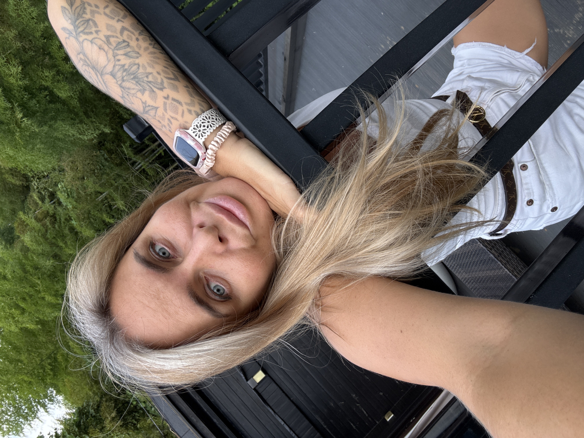 A blonde woman with striking blue eyes and fair skin leans her head on her hand, resting on a black metal railing. Her left arm, adorned with a floral tattoo and a pink smartwatch, is extended over the railing. She is wearing a white tank top and white denim shorts with a designer belt. Her blonde hair, streaked with grey, frames her face. The background features lush green foliage, suggesting an outdoor setting, possibly a garden or park. The scene appears to be captured in daylight, with soft lighting illuminating the woman's face. The location is not specified in the original image.