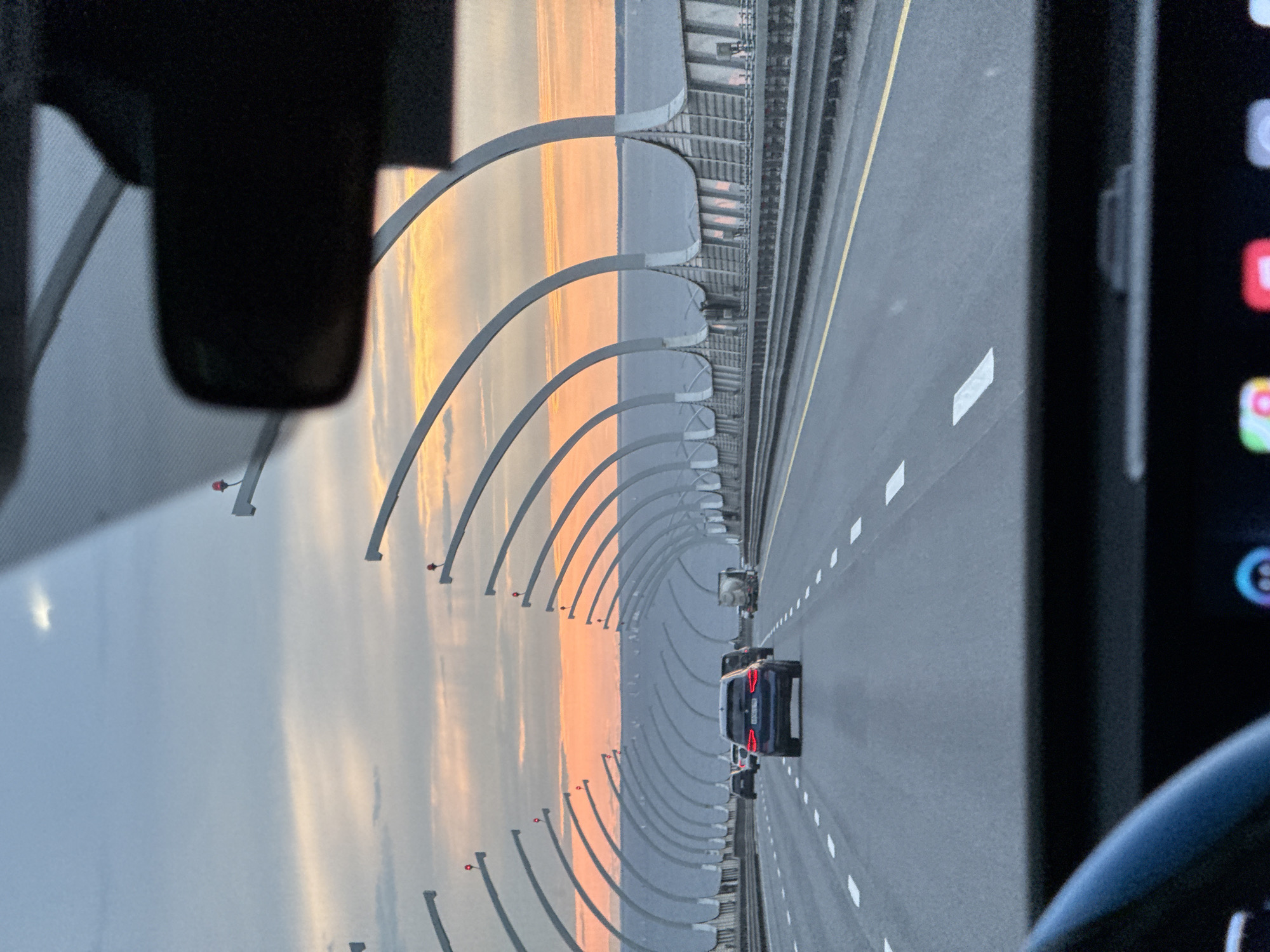 A forward-facing perspective from inside a vehicle shows a highway at sunset. The sky displays hues of orange and pink clouds. On either side of the road are large, arching structures that appear to be part of a sound barrier or canopy, with red lights visible on top. Several cars are driving on the highway, including a dark blue SUV in the foreground. The highway itself is paved, with white lane markings and a yellow line on the right. The water body to the right of the highway is calm and appears to be a bay or gulf. The image captures the KAD (Ring Road) in Saint Petersburg, Russia, specifically the section over the Gulf of Finland.