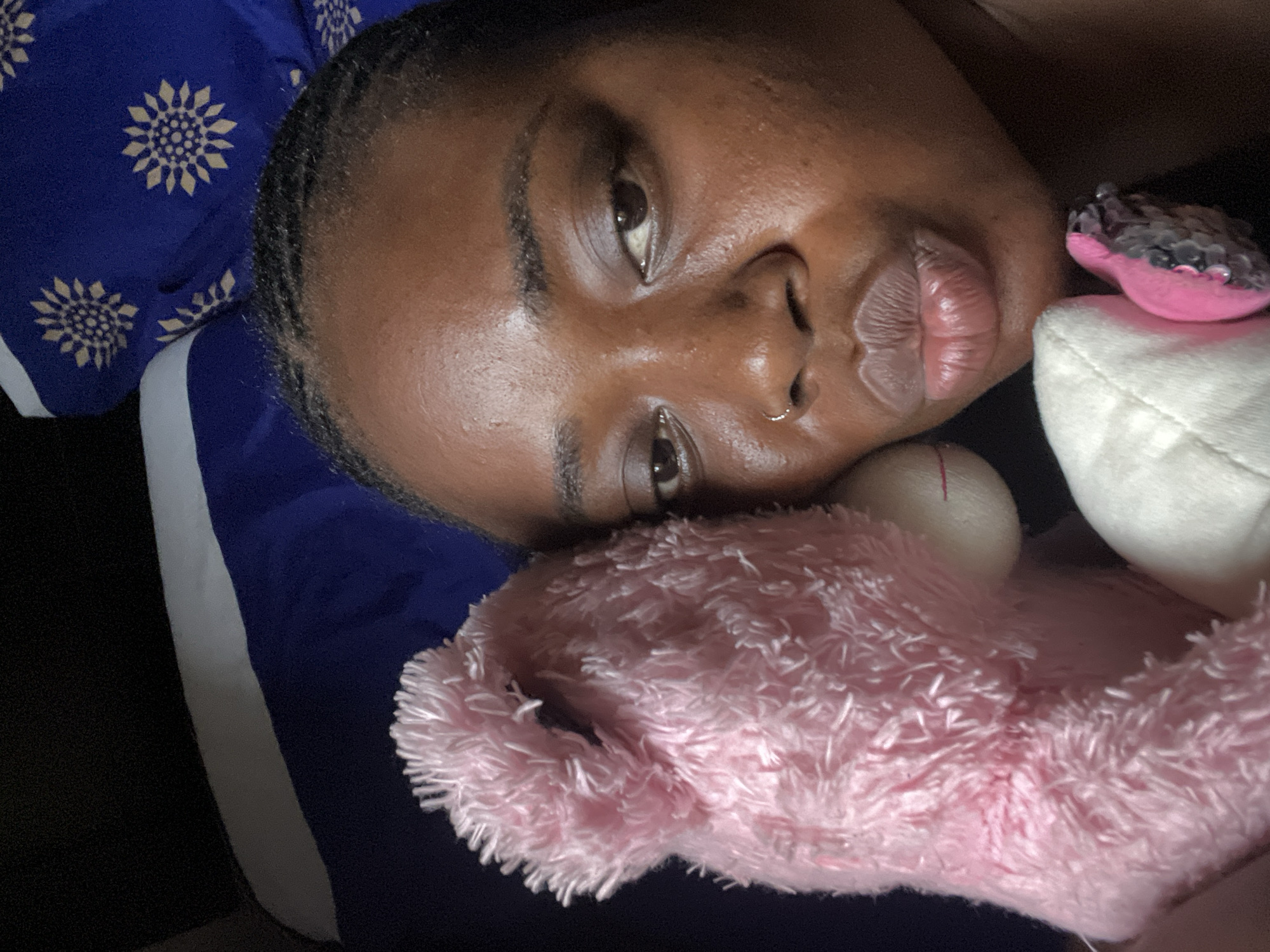 A person, likely female, with dark skin is captured in a close-up, slightly angled shot, lying down indoors. Her head rests on a vibrant blue pillow featuring a repeating pattern of golden, sunflower-like motifs. She has dark eyes, which look directly at the viewer, and full lips. A small gold hoop nose ring adorns her right nostril.
To her right, a soft, pink, fluffy object, resembling a stuffed animal or a plush blanket, is positioned close to her face. Further right, a small, bright pink item embellished with silver sequins sits on a white, possibly plush, surface. A blue and white striped fabric, potentially part of clothing or bedding, is visible along the left side of the frame. The overall scene suggests a moment of relaxation or rest in an indoor environment, likely a bedroom, in Abuja, Nigeria. The lighting is soft, providing no specific cues regarding the time of day. No text is visible in the image.