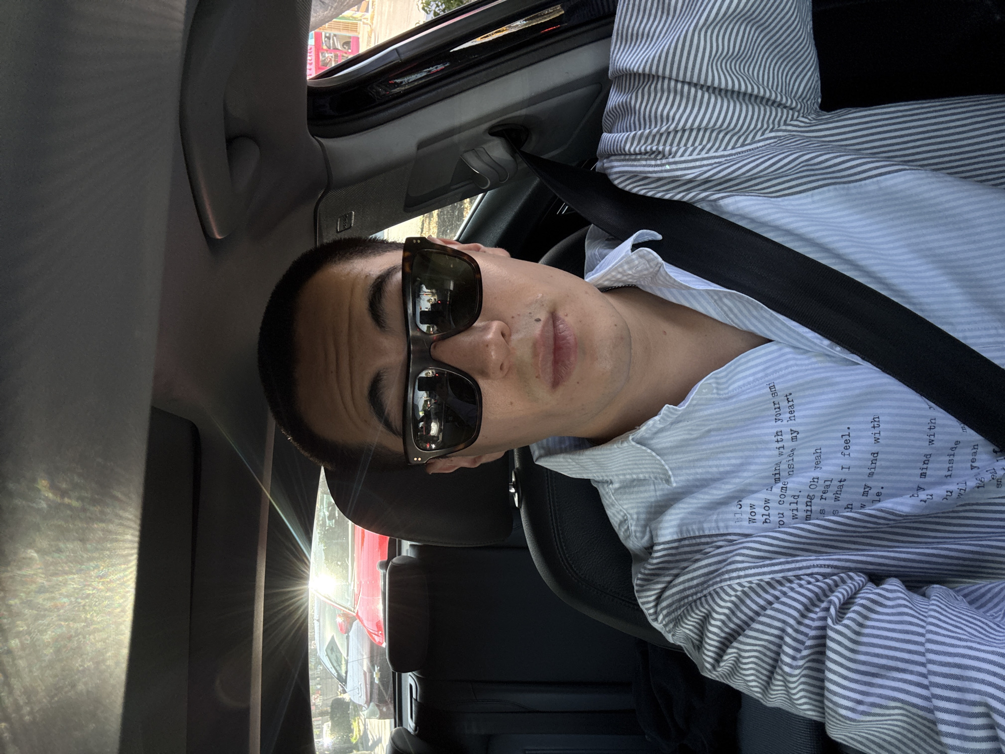 The image is a selfie captured from inside a car, originally rotated 90 degrees clockwise. It features a young adult male, likely of Asian descent, seen from the chest up. He has short, dark hair and wears dark, rectangular sunglasses with thick frames that obscure his eyes. Visible on his cheek and nose are some moles or freckles.
The individual is wearing a light blue and white striped button-up shirt. Prominent text printed on the shirt includes phrases such as "Wow blow mind with your smi," "you come inside my heart wild," "ming Oh yeah is real is what I feel," and "w my mind with." He is buckled into a black seatbelt.
The interior of the car is dark, with black seats and roof lining. A grab handle is visible above the passenger side window. Bright sunlight pours into the car from the right side, creating significant glare, lens flare, and a rainbow-like diffraction pattern on the car's interior fabric. This indicates a sunny daytime setting. Reflections in the sunglasses show parts of the exterior environment, including other vehicles (a red car is clearly visible) and buildings, suggesting an urban or suburban street scene. The person appears to be casually posing for the photograph. This scene takes place in Puebla, Mexico.
