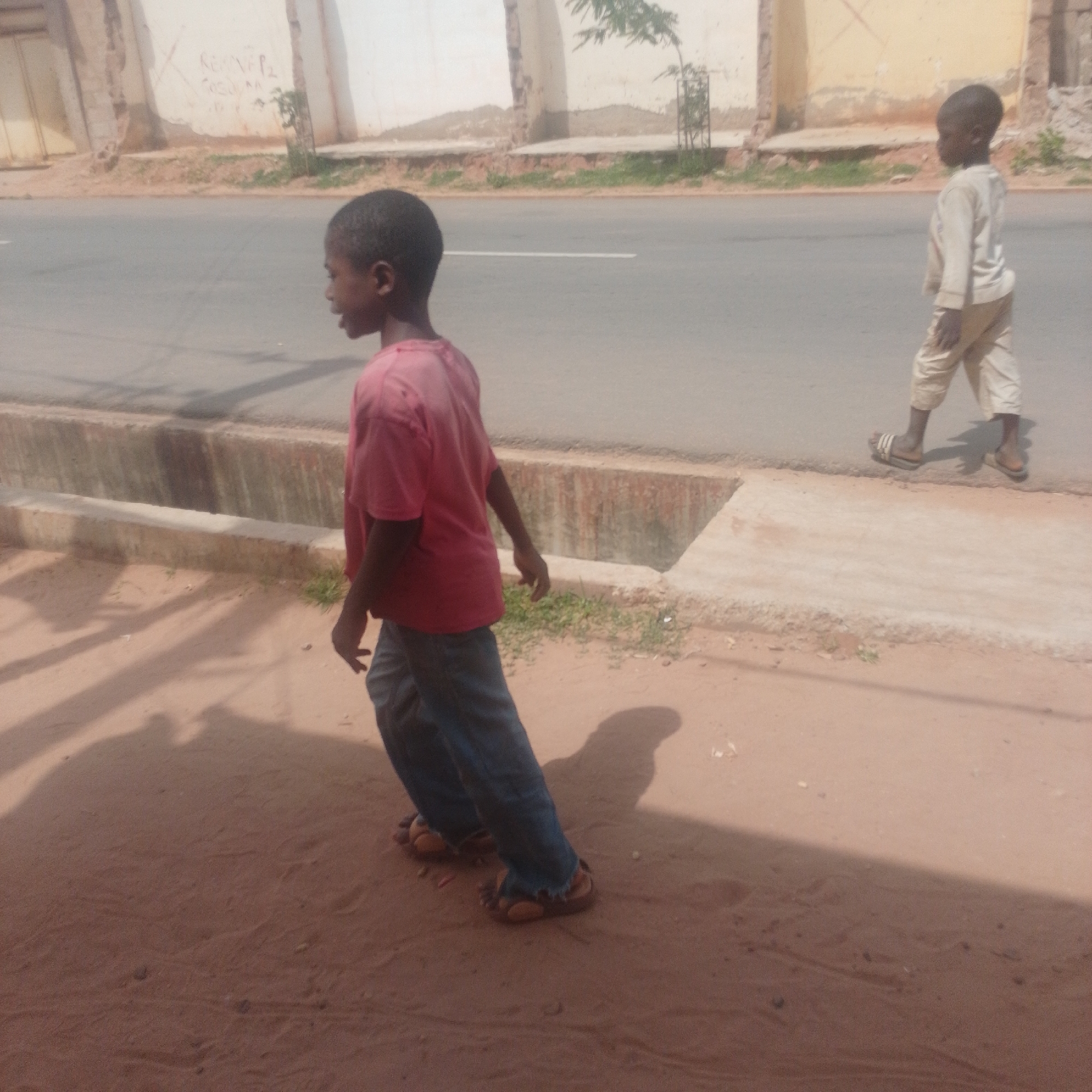 The image captures two young boys walking along a dusty roadside in Gombe, Nigeria. The boy in the foreground, facing left, is wearing a pink t-shirt and blue jeans. His body is angled away from the camera, and he appears to be walking or running. The second boy, further down the road and closer to the street, is walking away from the camera, facing right. He is dressed in a long-sleeved cream-colored shirt and light-colored pants. Both boys are wearing sandals.
The setting is a street with a paved road to the right and a dirt path with some sparse vegetation on the left. A concrete drainage channel runs along the edge of the road. In the background, there are buildings with weathered walls, some displaying graffiti. The lighting suggests it is daytime, with strong shadows indicating the sun is present, possibly mid-morning or afternoon. The overall atmosphere is casual, with the children engaged in their daily activities. There is no discernible text within the image itself.