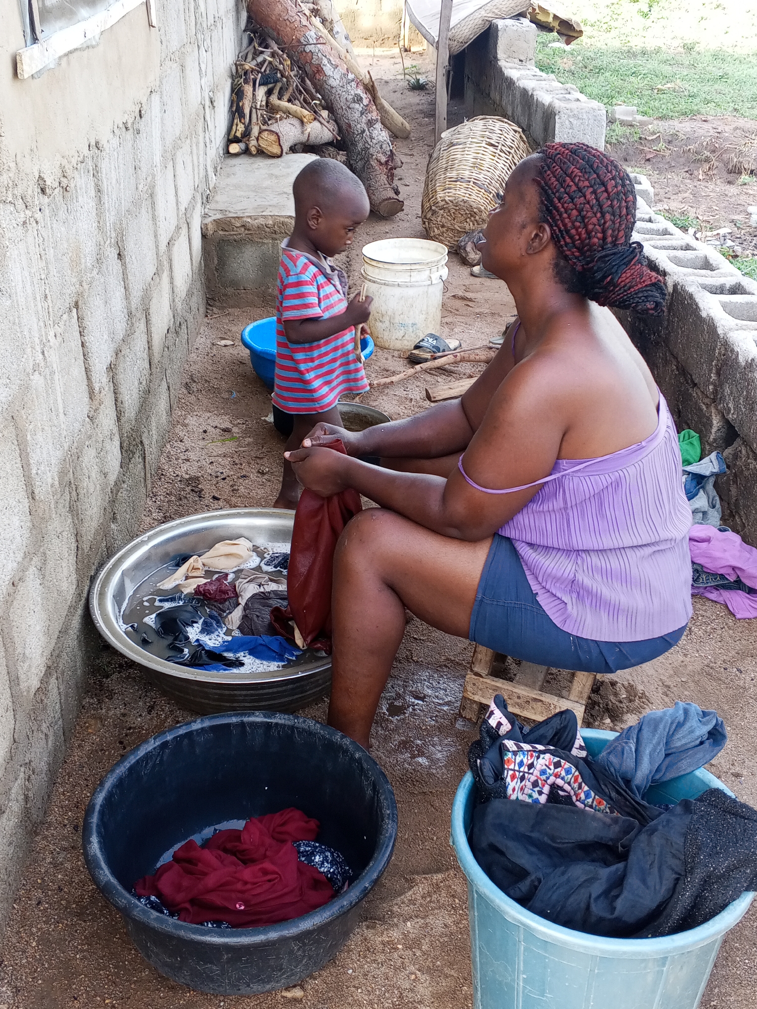 This photograph captures an outdoor scene in Adonkolo, Nigeria, depicting a woman hand-washing clothes while a young child stands nearby.
The main subject is a woman with dark skin and braided hair, styled in red and black sections pulled into a bun. She is seated on a low wooden stool, wearing a light purple tank top and dark shorts. Her right shoulder strap is slightly slipped down. She is actively engaged in washing, holding a reddish garment over a large silver basin filled with soapy water and other clothes. Her gaze is directed upwards and to the right, suggesting she is looking at something or someone just out of frame, possibly the child.
Beside her, a young child with short hair stands, facing slightly away from the camera. The child wears a red and blue striped t-shirt and dark shorts, and appears to be holding a small stick or object, looking down at it.
Surrounding the woman are several containers related to laundry: the large silver basin for washing, a black plastic basin on the ground holding wet clothes, and a light blue plastic bucket containing what appears to be dry or dirty clothes. A small blue plastic bowl is also visible near the child's feet.
The setting is an unpaved area with sandy dirt ground, suggesting a backyard or open space next to a residence. To the left, an unfinished concrete block wall forms part of a building structure. In the background, logs and cut wood are stacked against a raised concrete platform, likely for firewood. A wicker basket and a white plastic bucket are also present in the mid-ground. Further back, green foliage indicates an outdoor, somewhat natural environment. The lighting suggests it is daytime, likely during morning or late afternoon, with clear weather. No visible text is present in the image. The scene portrays a moment of daily domestic activity.