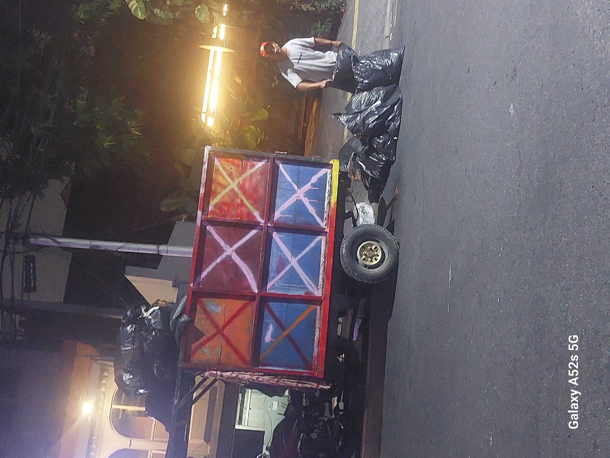 The image shows a street scene in South Jakarta, Indonesia at night. A man wearing a white t-shirt and a red cap is standing on the sidewalk next to a small truck loaded with trash bags. He appears to be holding one of the trash bags, suggesting he is in the process of loading or unloading it. The truck has a colorful, painted cargo bed with red, orange, and blue sections, each divided by white X markings. Several large black trash bags are piled on the ground next to the truck. In the background, there is a building with arched windows and some foliage. The street is paved, and there are streetlights visible, indicating an urban environment. The overall impression is a moment captured during waste collection activities.